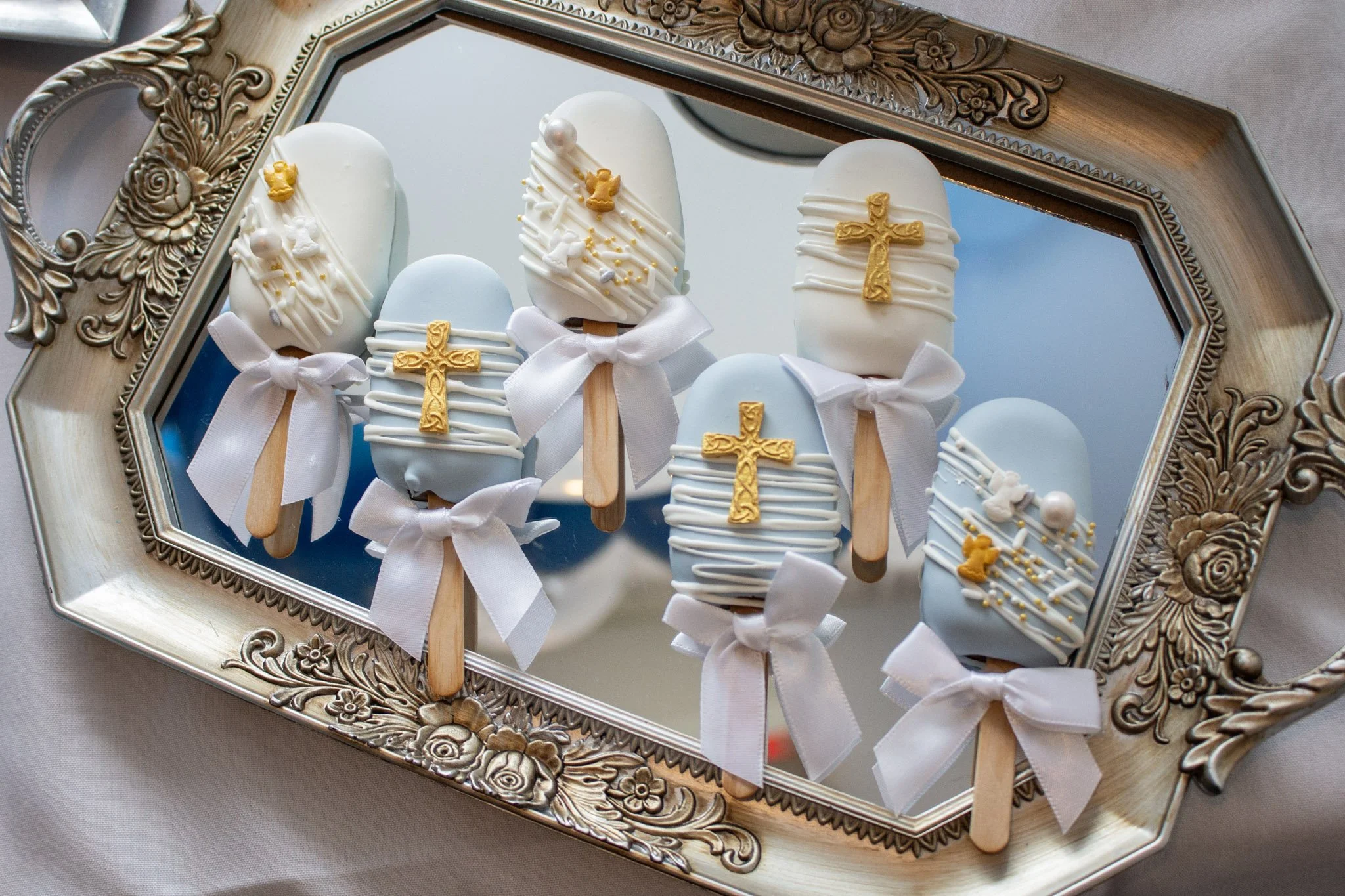 Ice cream popsicles decorated with white and light blue icing, gold crosses, white bows, and small gold and white edible decorations, displayed on a decorative ornate silver tray.
