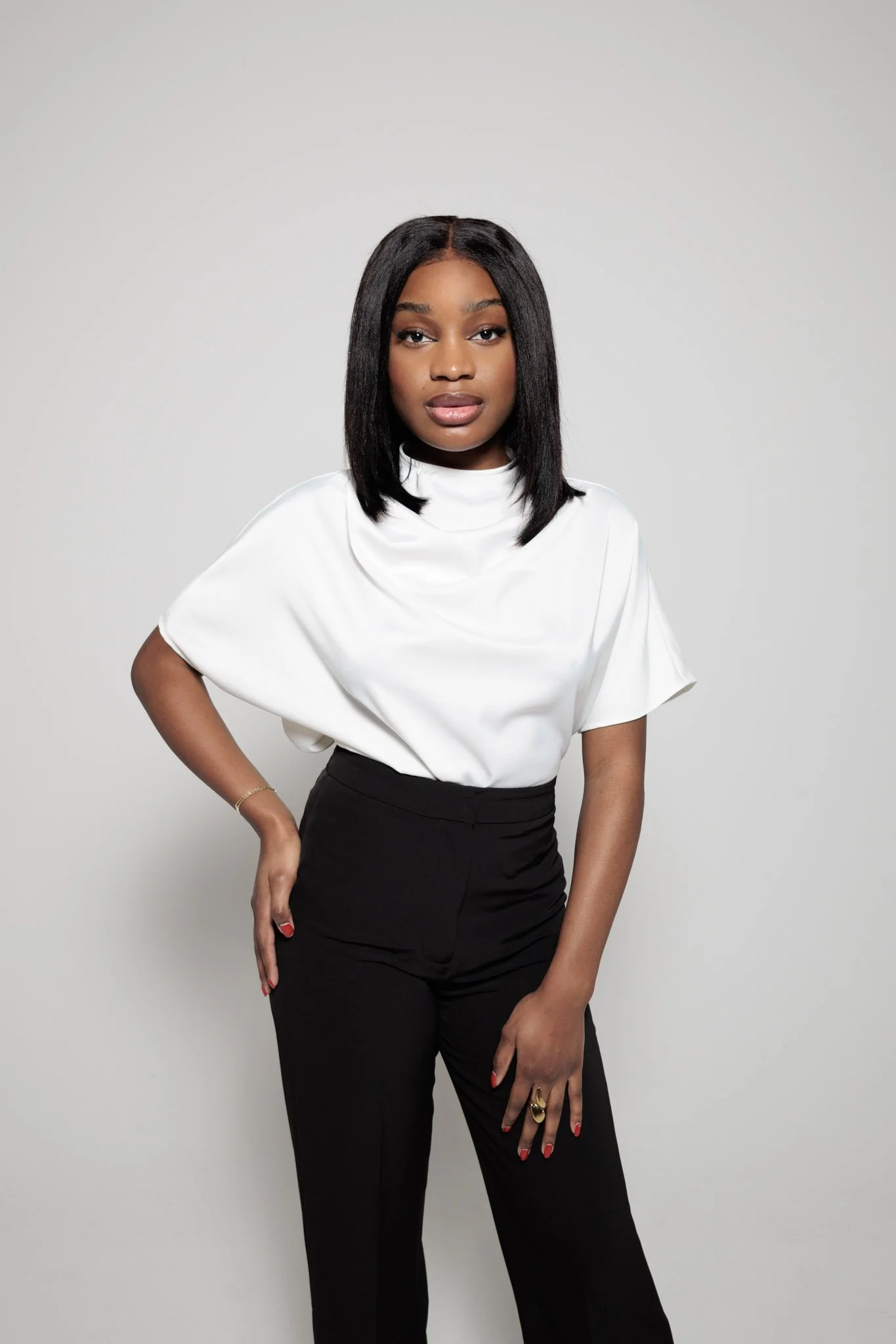 A young woman with dark hair, wearing a white top and black pants, standing against a plain background.