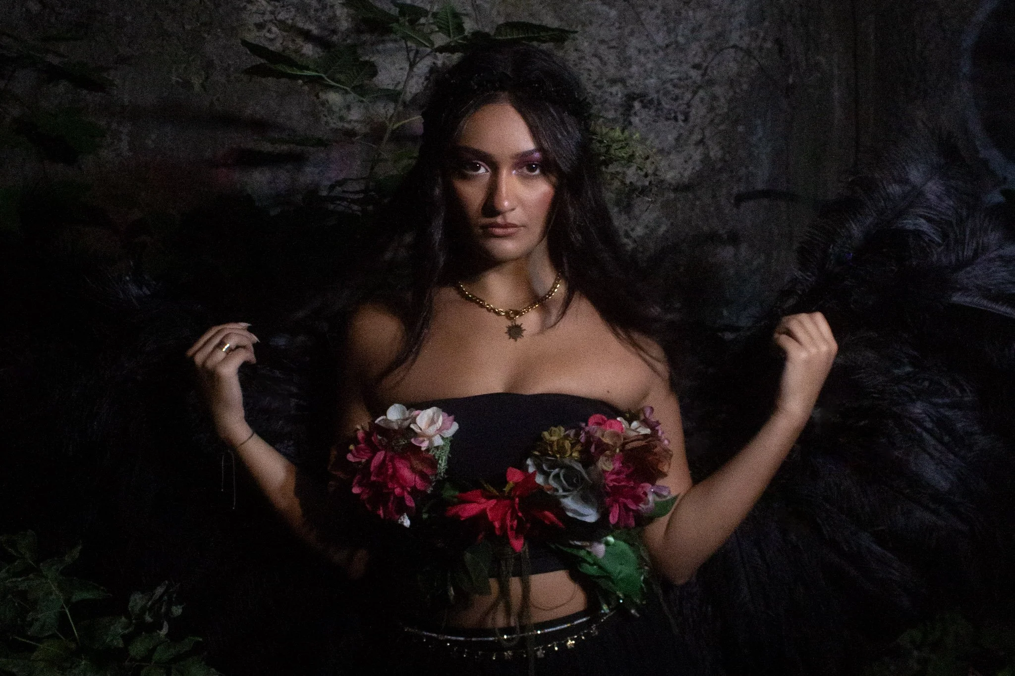 A woman with long dark hair and dark clothing, lying against a rocky background, surrounded by foliage, with feathered wings and wearing jewelry, looking directly at the camera.