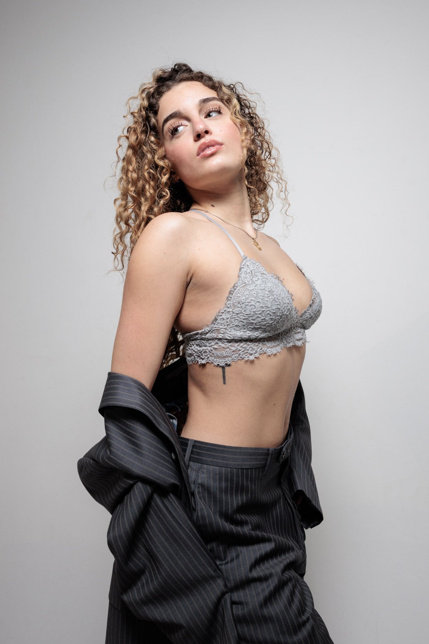 A young woman with curly hair poses against a plain background, wearing a gray lace bralette, black pinstripe pants, and a dark blazer draped off her shoulders.