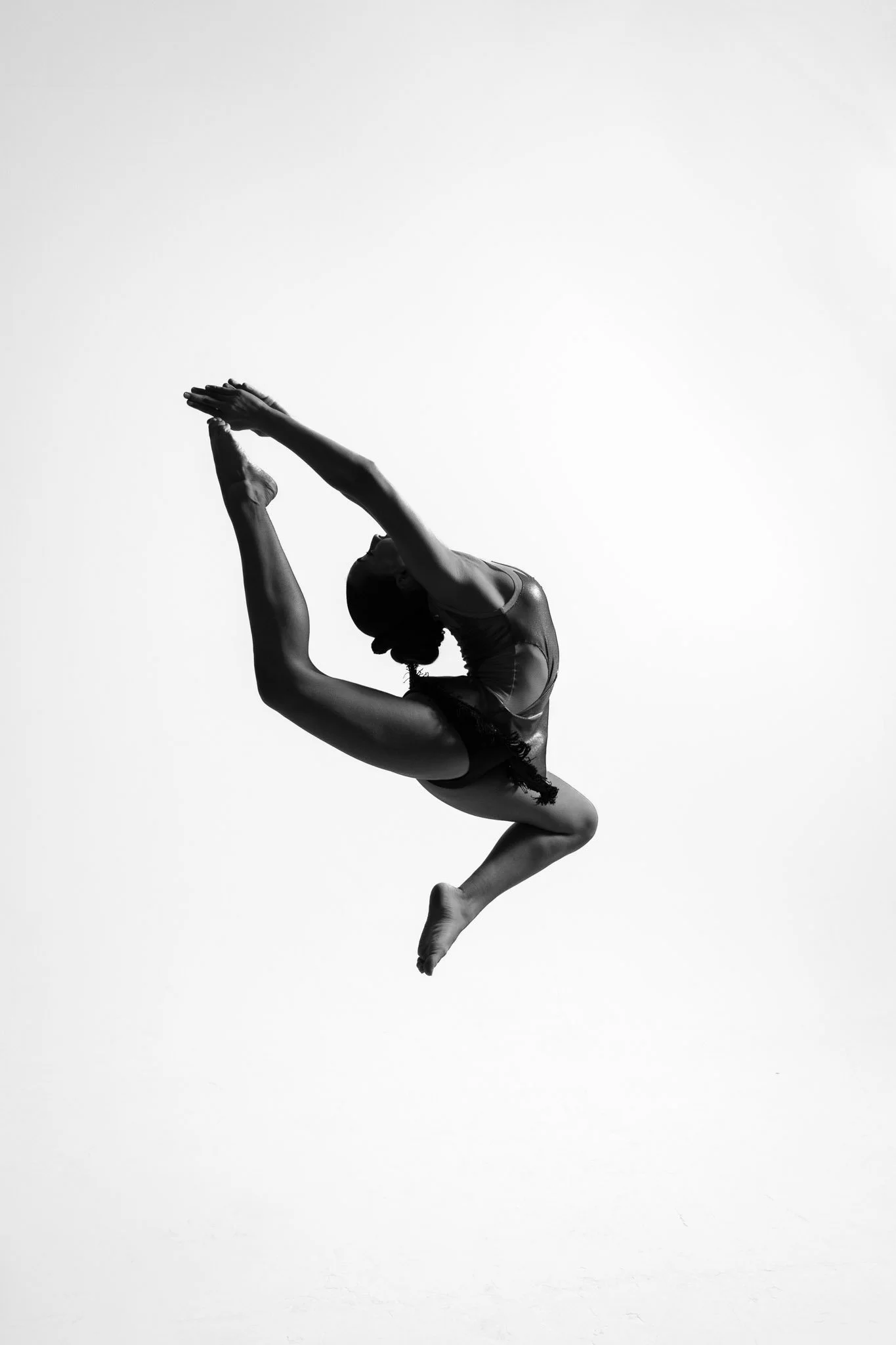 A black and white photo of a female gymnast in a backbend pose with arms extended overhead, standing on one foot.