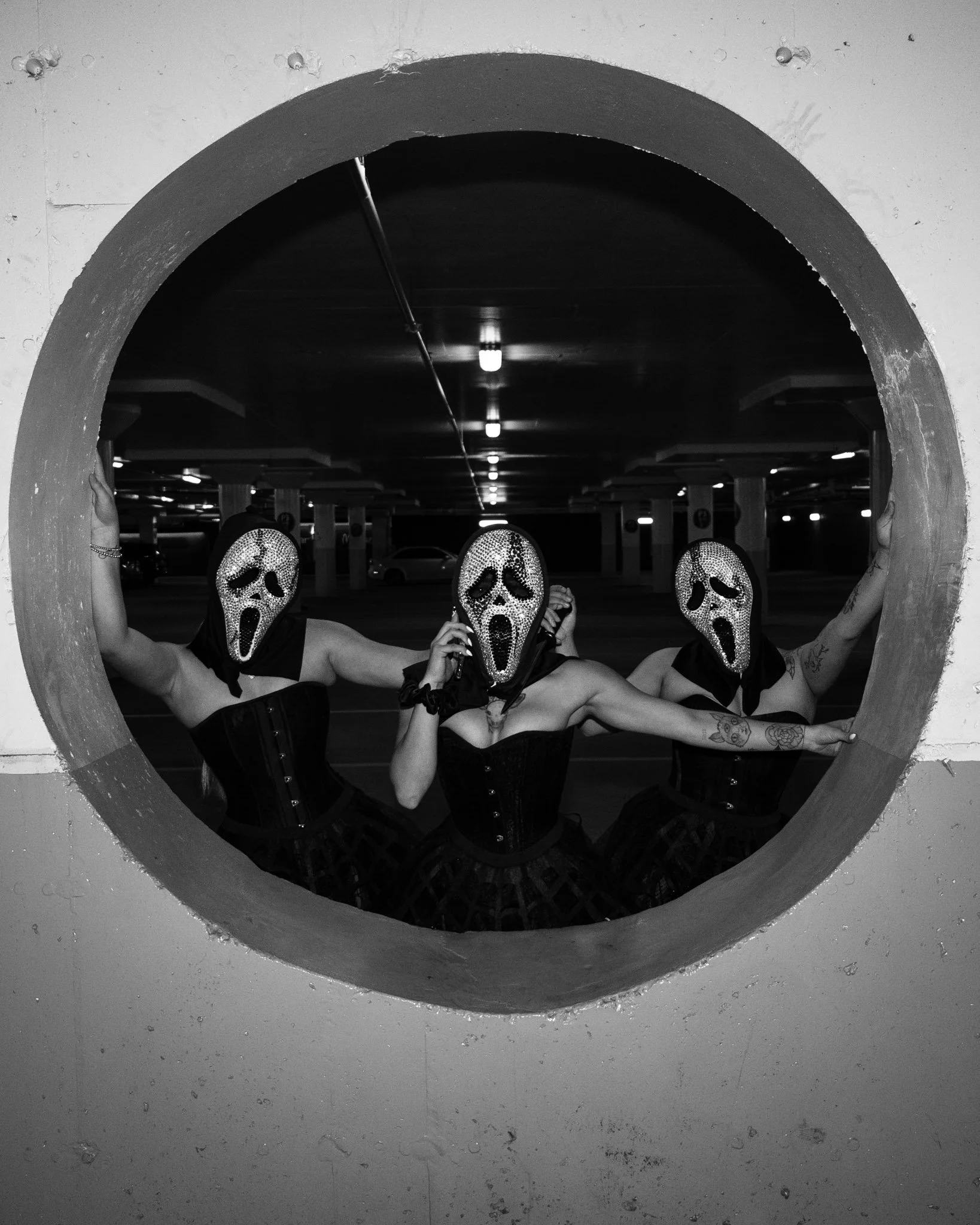 Three people wearing masks with the scream face from the movie Scream, dressed in black corsets and skirts, posing in a parking garage seen through a circular opening.
