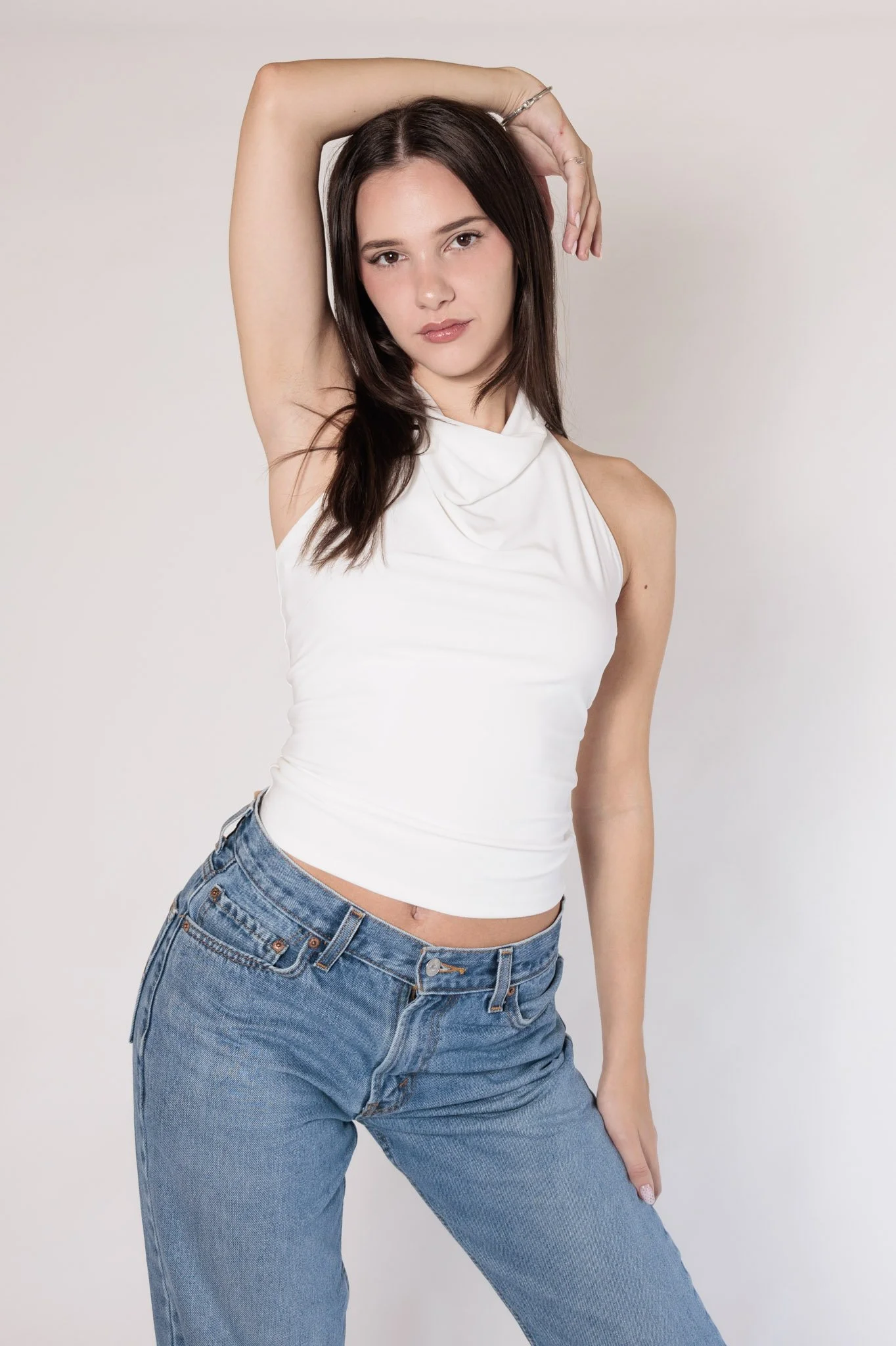 Young woman with dark hair in a white sleeveless top and blue jeans posing with one arm raised behind her head against a plain white background.