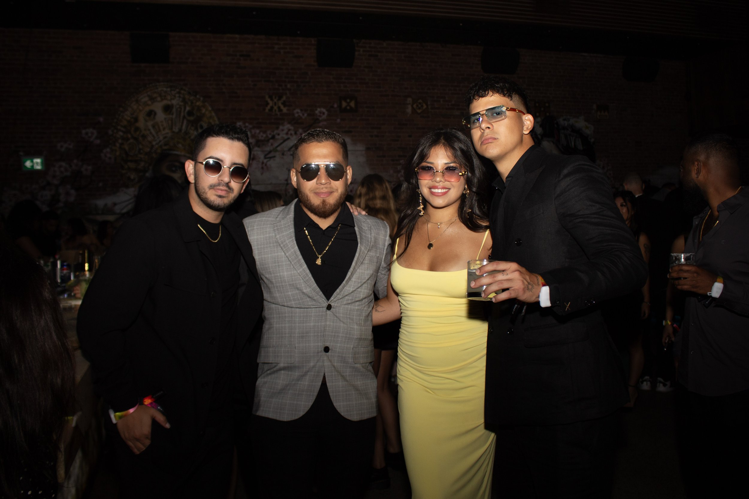 Group of five young adults dressed in formal attire, four men and one woman, at a party or nightclub, with some holding drinks and wearing sunglasses.