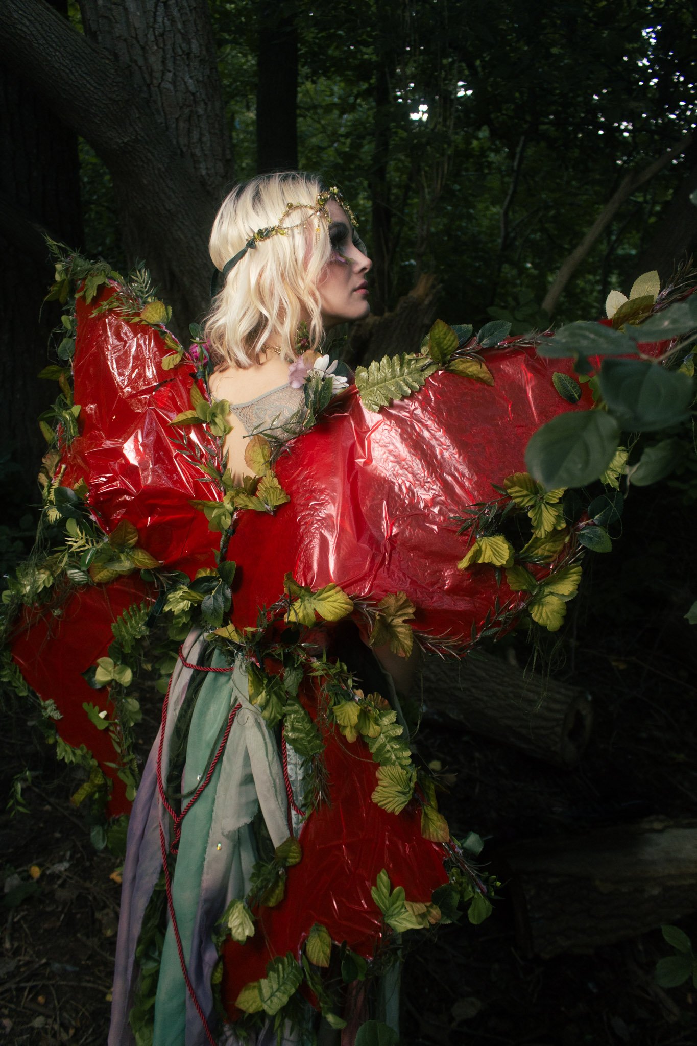 A woman with blonde hair in a forest, dressed in a fantastical costume with large red and green leaf-like elements and a gold tiara.