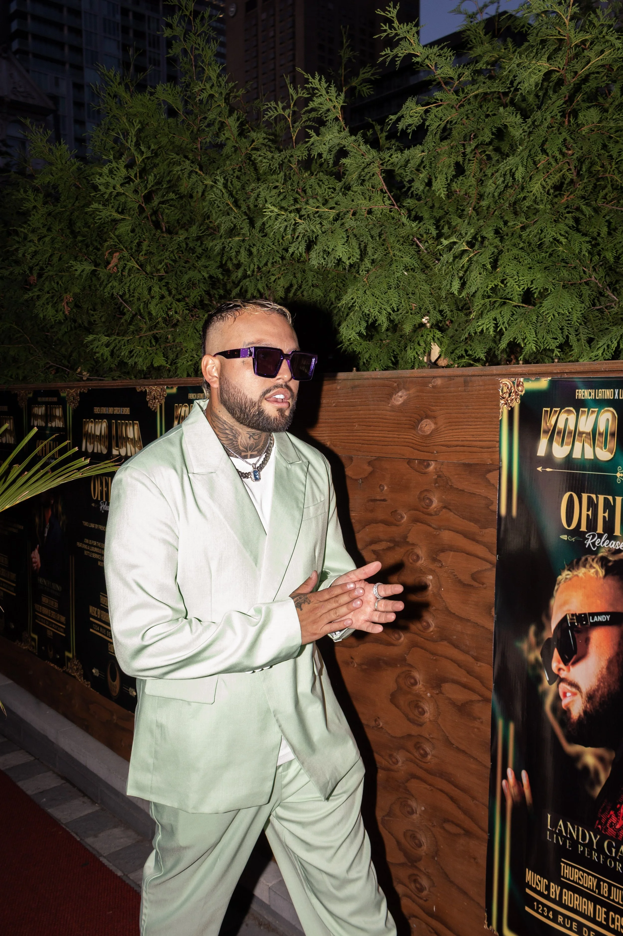 A man with tattoos, wearing sunglasses and a light green suit, stands in front of a wooden wall with posters for a music event, with greenery and buildings in the background. Montreal latino artist singer event at Yoko Luna. Landy Garcia. 