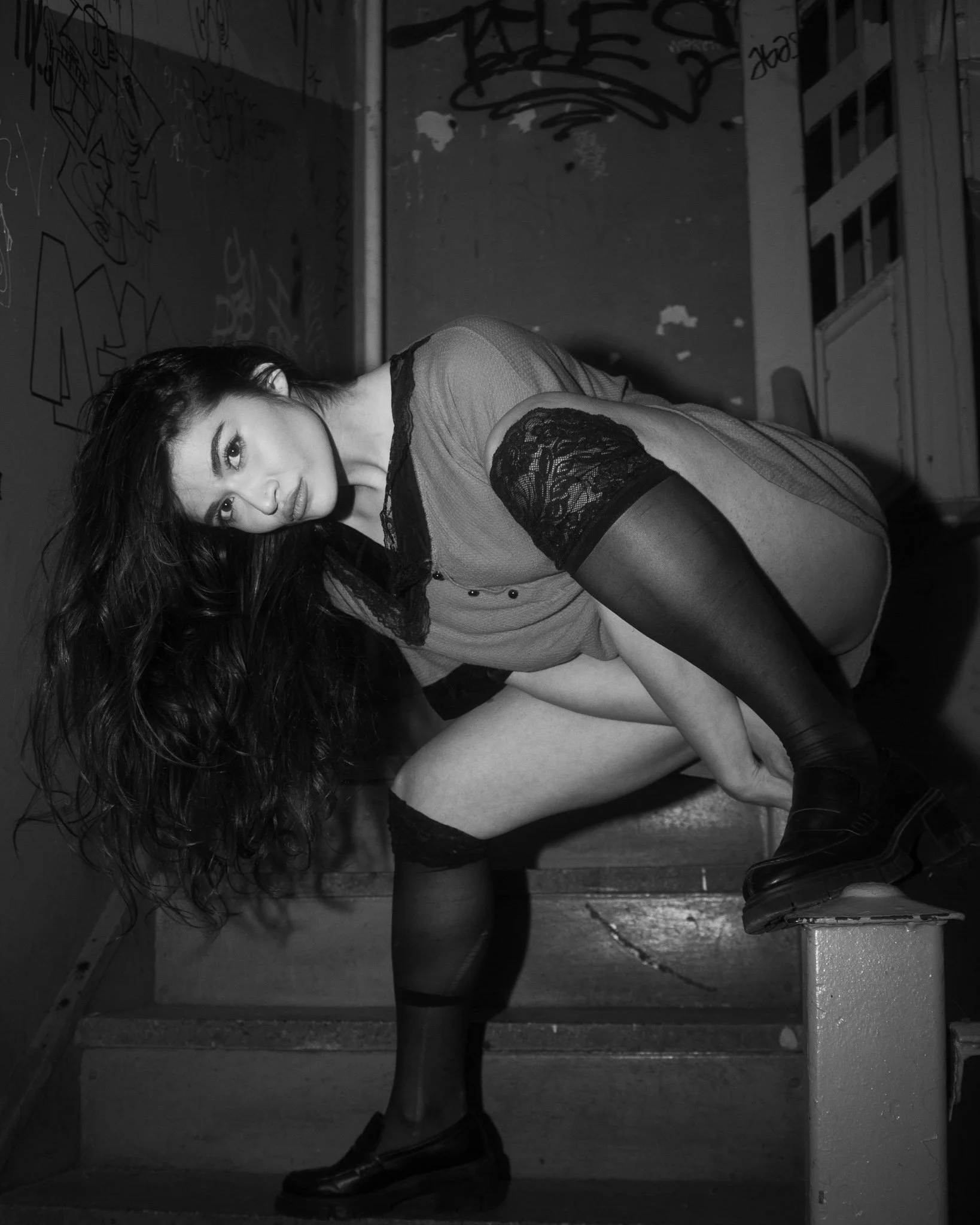 A woman with long dark hair and light skin is crouching on stairs in an industrial or urban setting with graffiti on the wall behind her. She is wearing a loose top, lace thigh-high stockings, and shoes, looking directly at the camera.