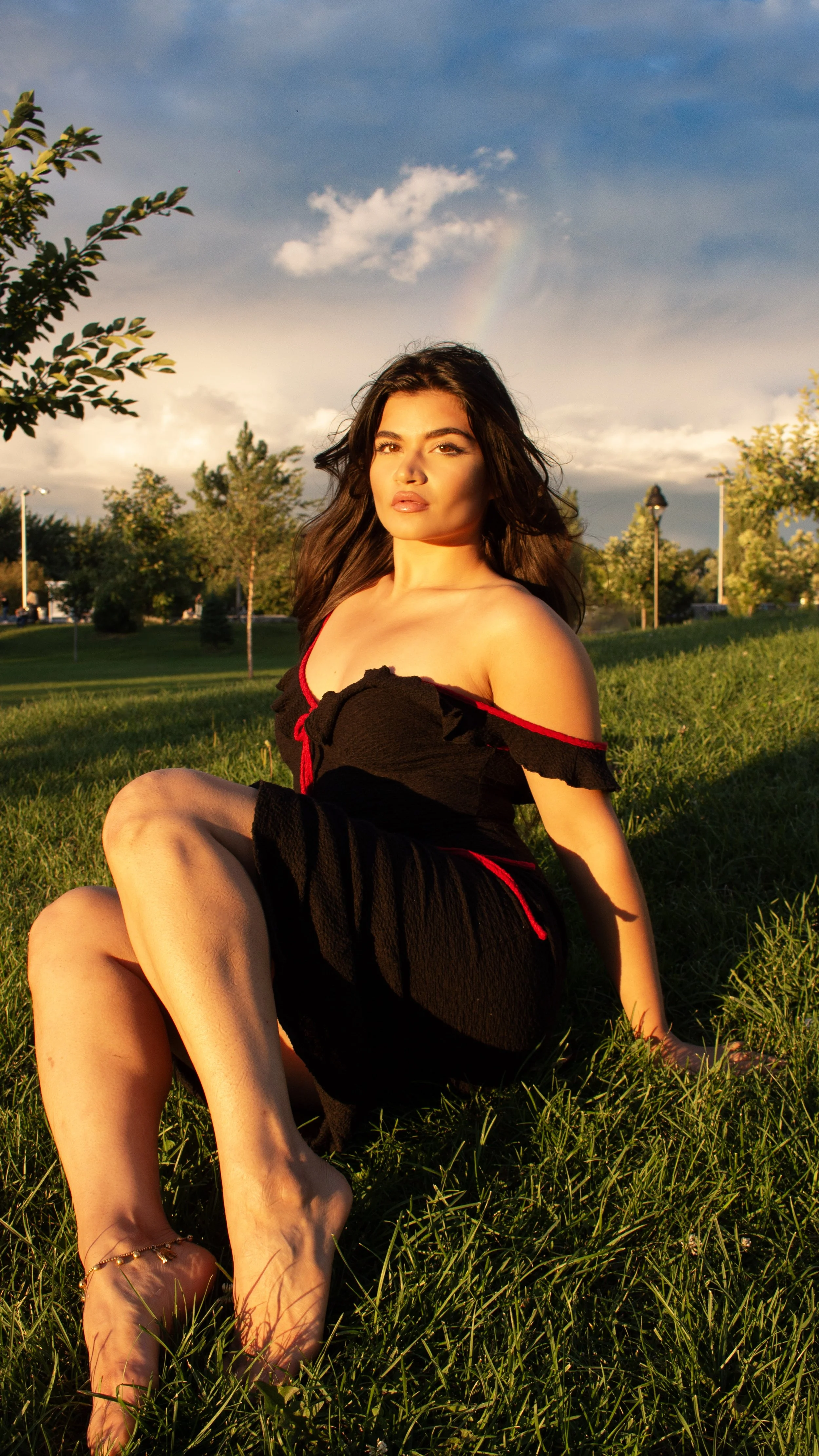 A woman with long dark hair sits on grass in a park during sunset, wearing a black off-shoulder dress with red accents, with a rainbow visible in the cloudy sky behind her.