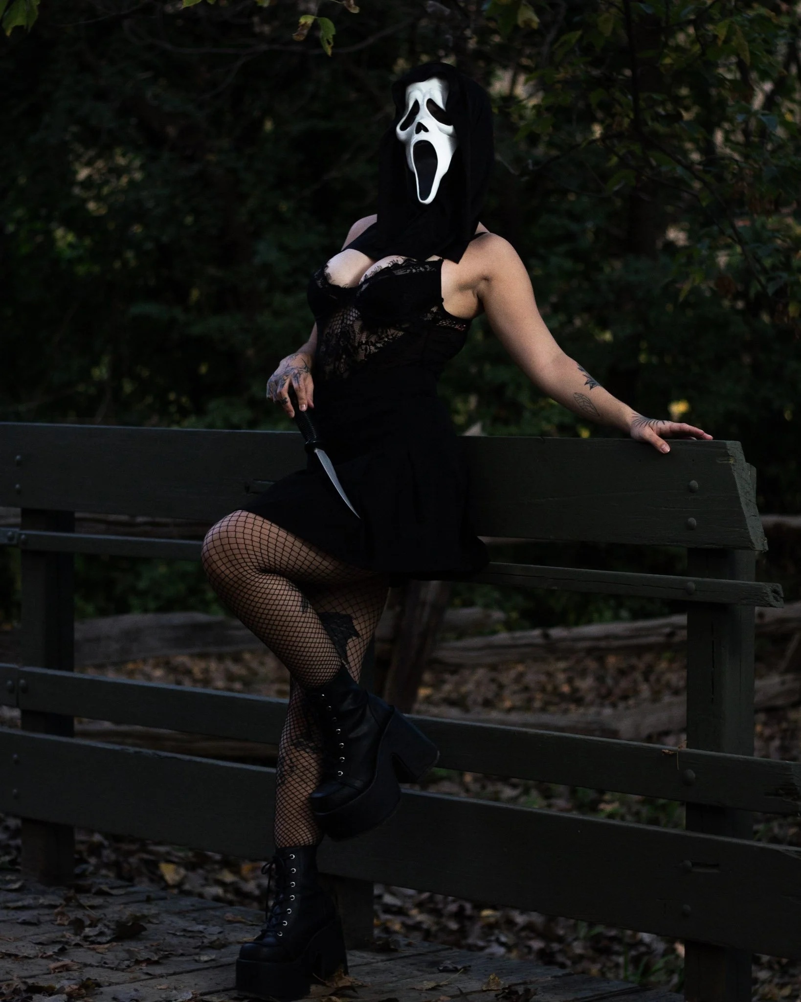 Person sitting on a park bench wearing a black lace dress, fishnet stockings, black platform boots, holding a large knife, and wearing a ghost face mask with a black hood over their head, in a wooded area.