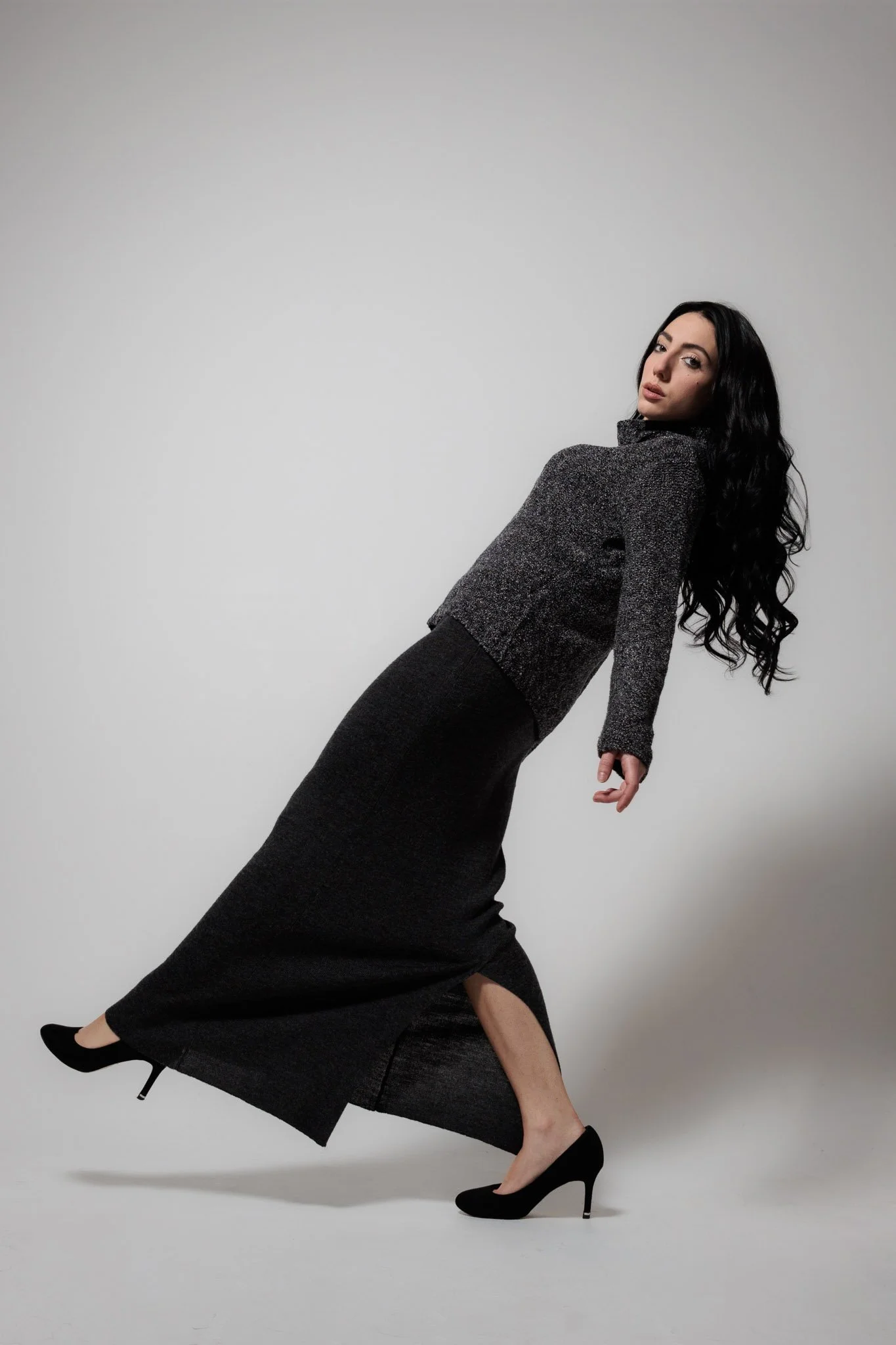 A woman with long black curly hair wearing a dark grey sweater and a long black skirt with a slit, posing against a plain background.