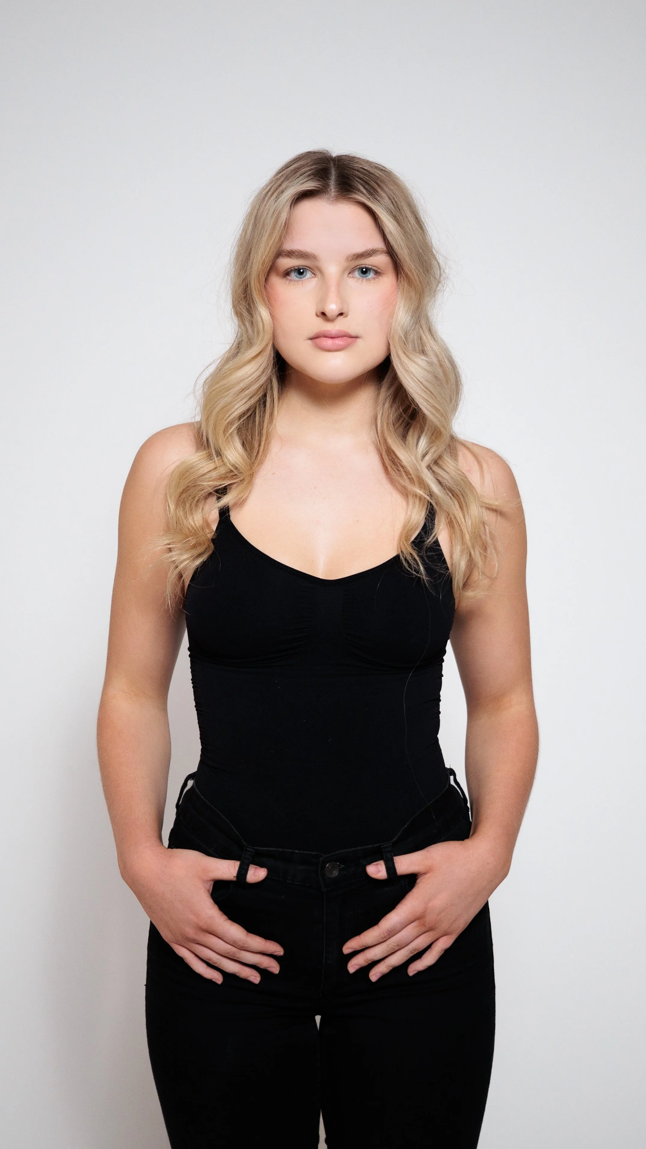 A young woman with blonde wavy hair, blue eyes, and fair skin, wearing a black sleeveless top and black pants, standing against a white background.