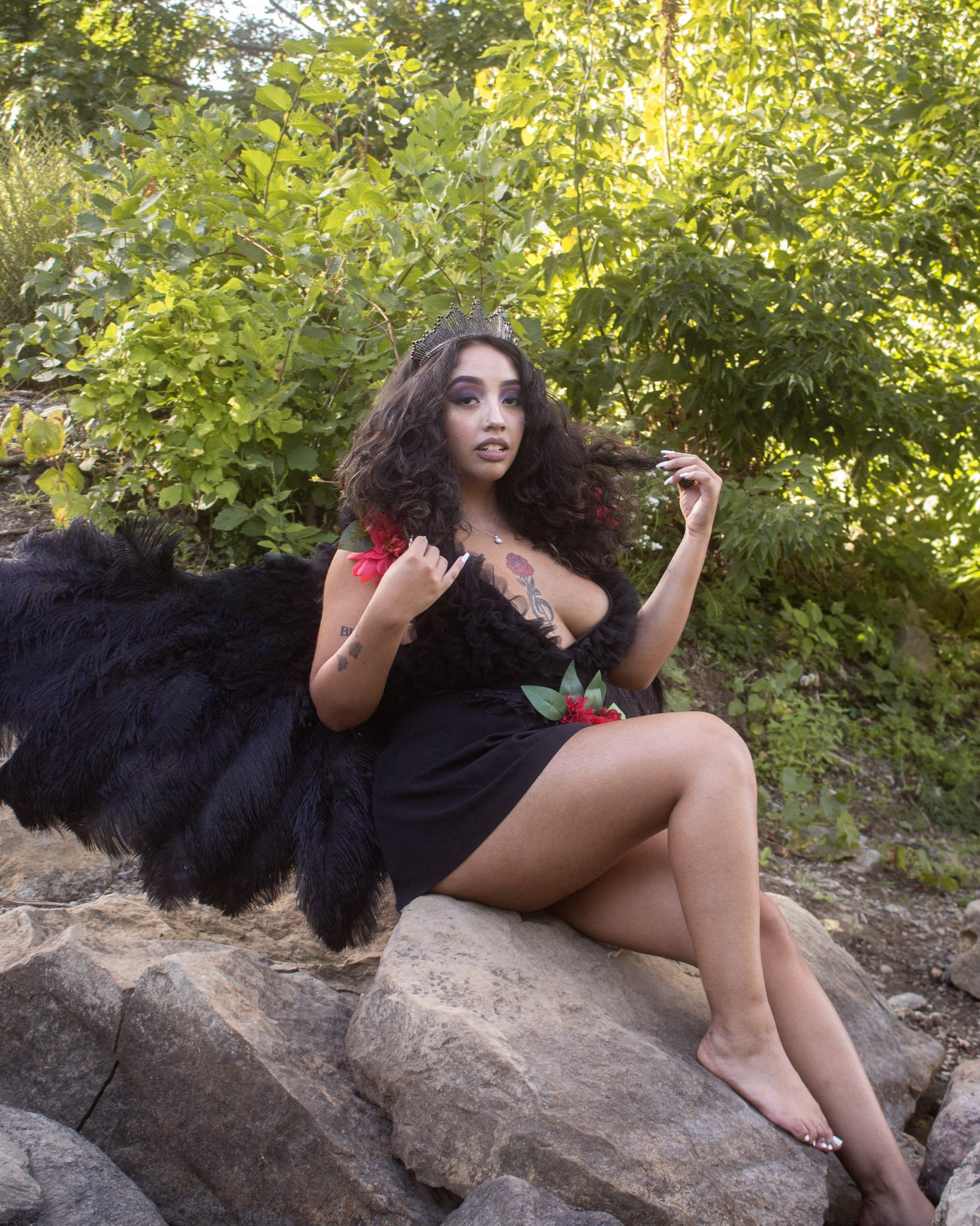 A woman with dark curly hair and a crown, sitting on rocks outdoors surrounded by green foliage, wearing a black dress with red flowers, and black wings, posing for the camera.