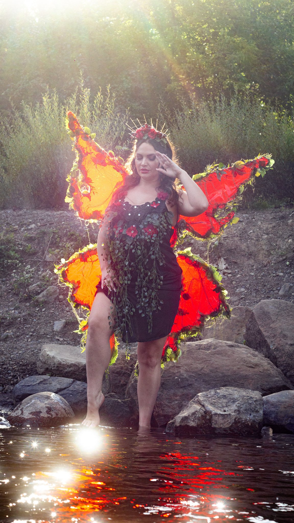A woman dressed as a fairy or mythical creature stands in a shallow creek or river, surrounded by rocks and the natural landscape, with lush green bushes and trees in the background. She is wearing a black dress with floral decorations, has large, vi