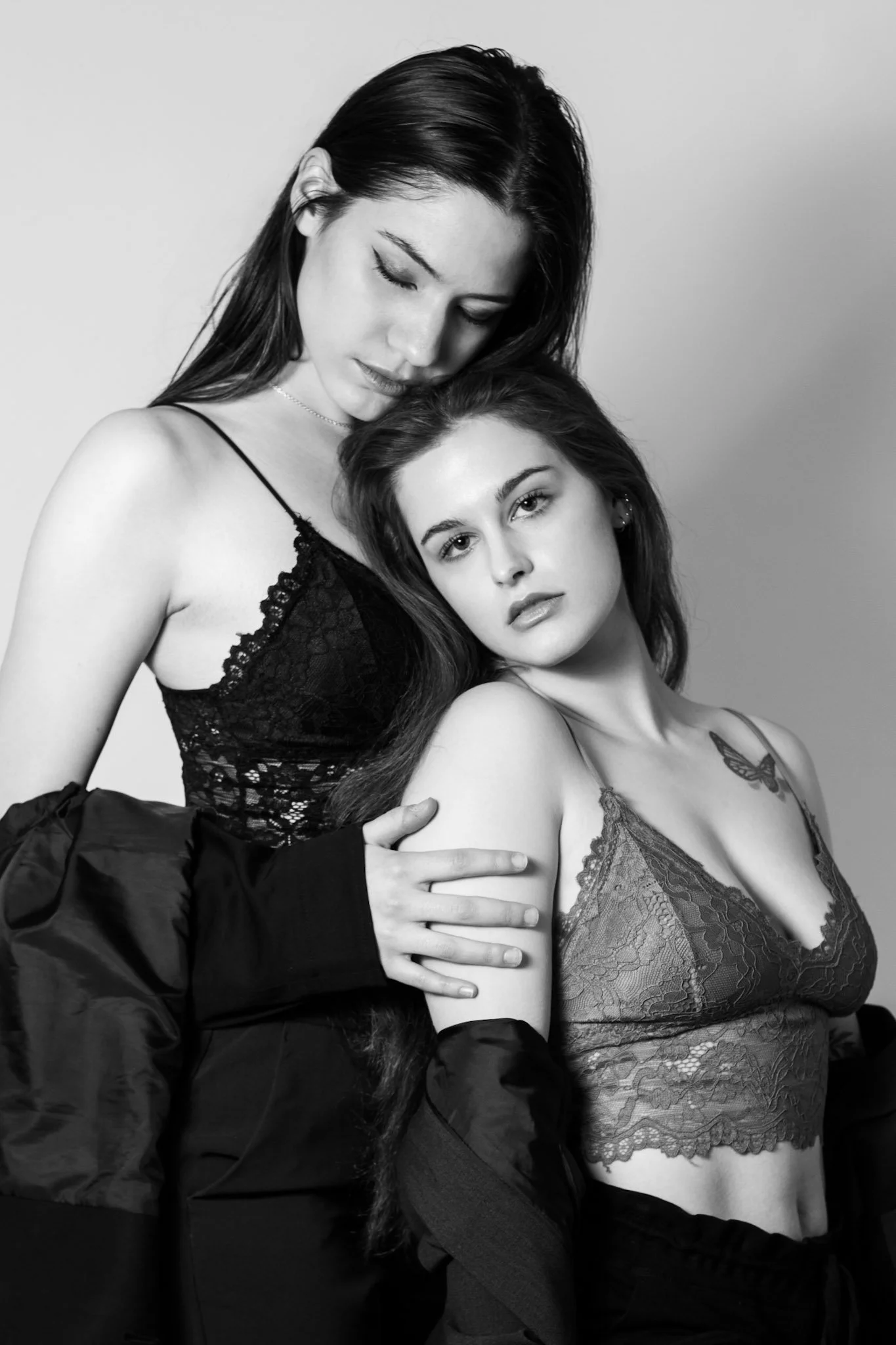Two women wearing lace lingerie and black jackets, posing closely together against a plain background. One woman has dark hair and the other has lighter hair, with a butterfly tattoo visible on her chest.