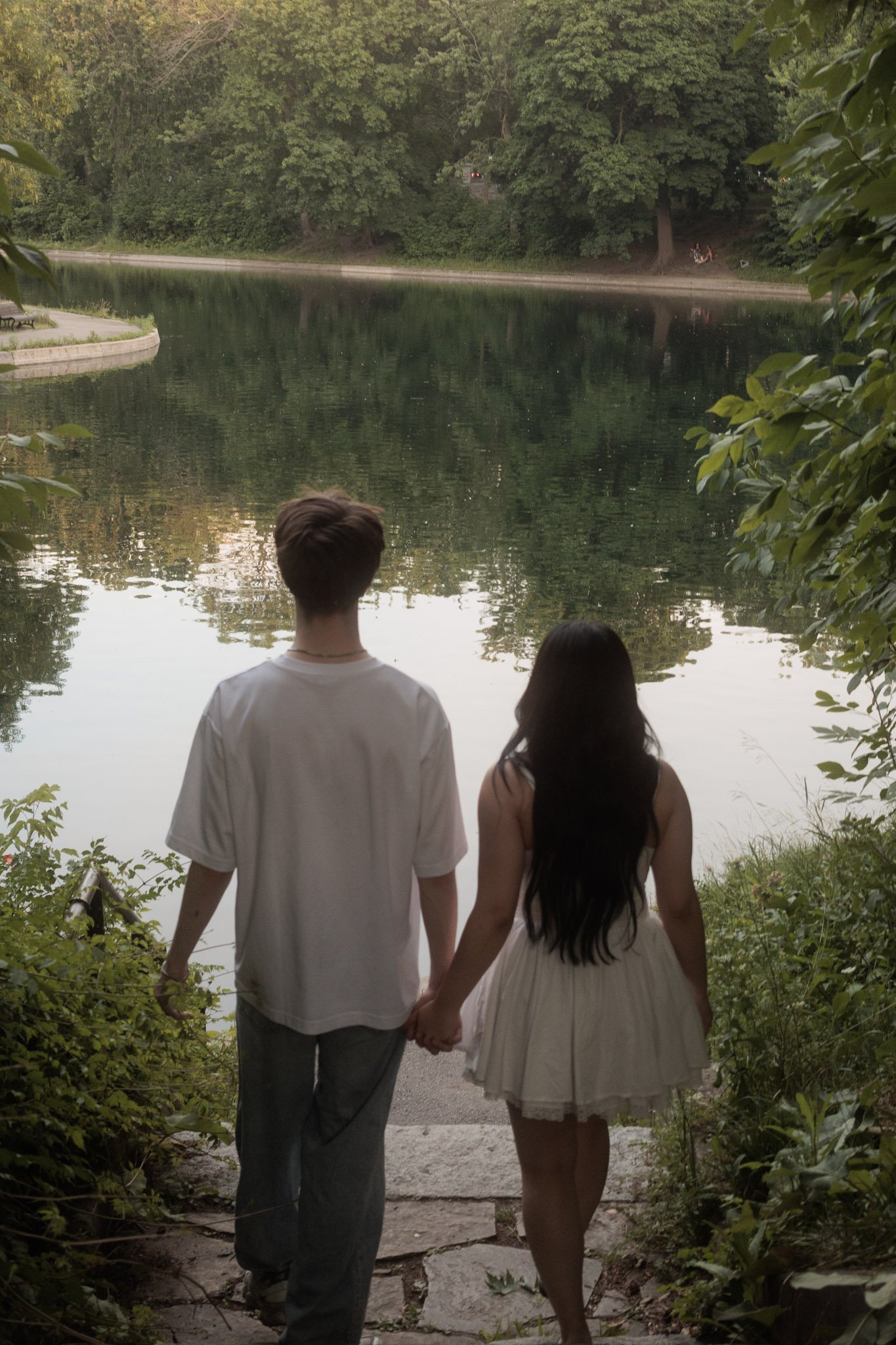 A young couple is holding hands and walking down steps towards a calm lake surrounded by greenery, with trees reflected on the water.