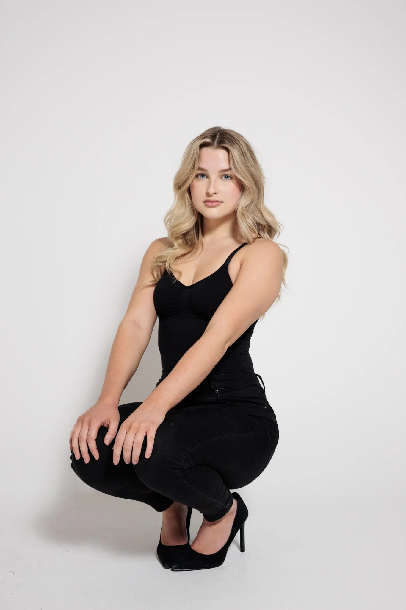 A woman with long, wavy blonde hair is squatting on the floor against a white background. She is wearing a black spaghetti strap top, black jeans, and black high heels.