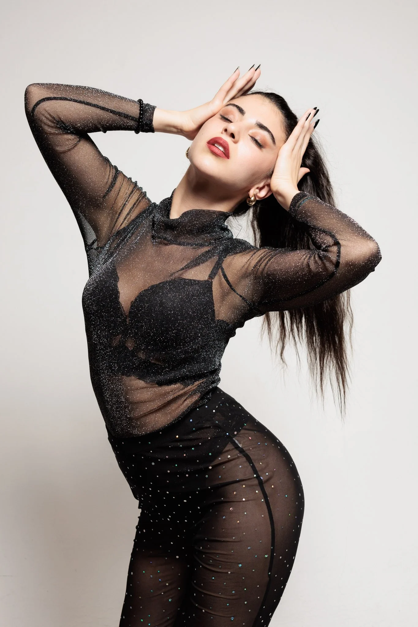 A woman with long dark hair and red lipstick, wearing a sheer black, glittery top and matching pants, poses with her eyes closed, hands on her head, and an expressive look against a plain background.