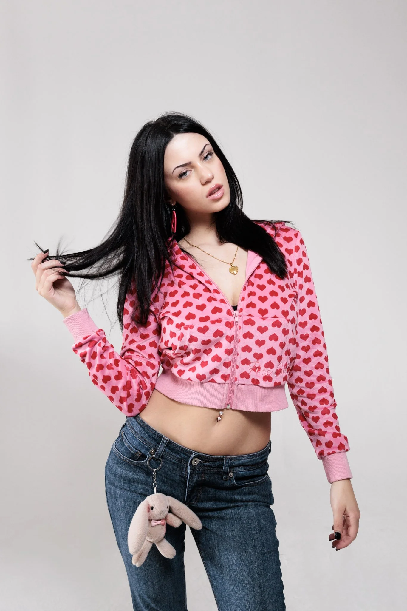 A young woman with long black hair, wearing a pink zip-up jacket with red heart patterns, blue jeans, and a bunny plush keychain attached to her pocket, standing against a plain light gray background.