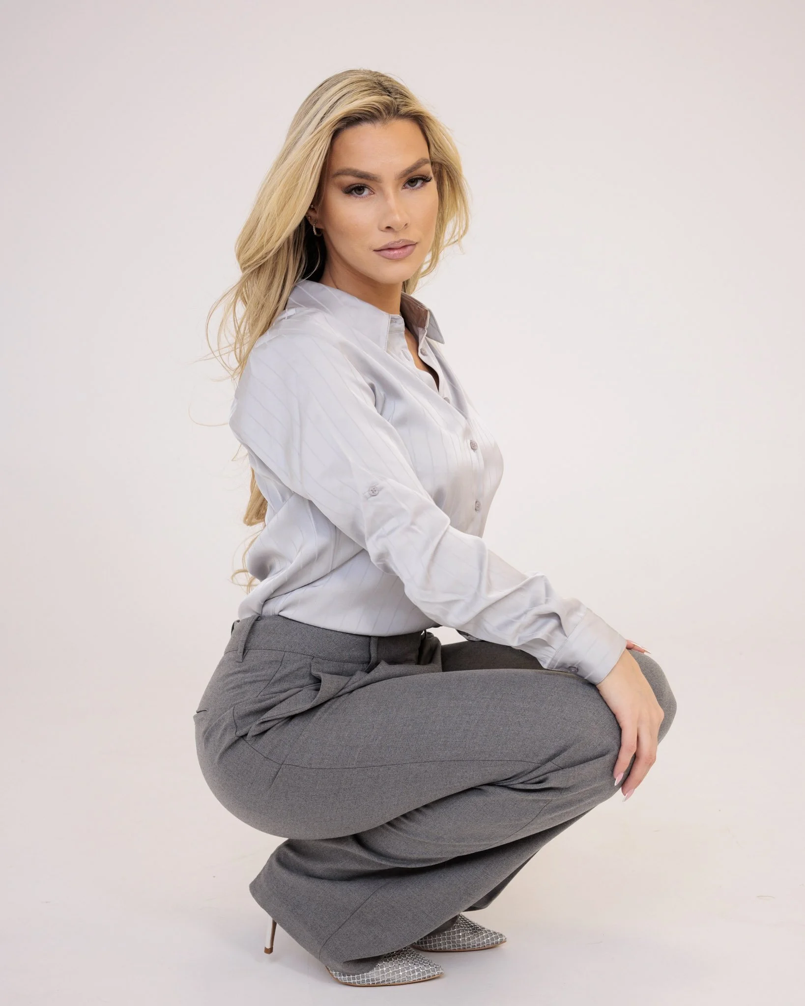 A woman with long blonde hair, wearing a light-colored striped button-up shirt and gray dress pants, is crouching with one knee bent and looking into the camera. She is posing against a plain white background.