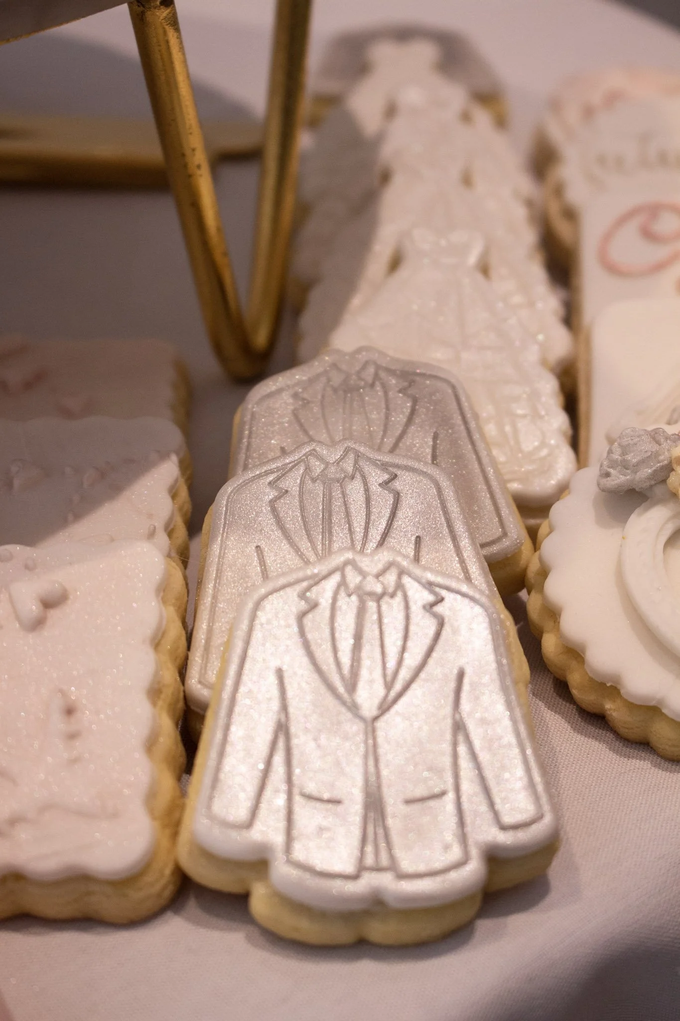 Cookies decorated with white icing, featuring a tuxedo suit outline. Other cookies in the background are shaped and decorated for a wedding celebration.