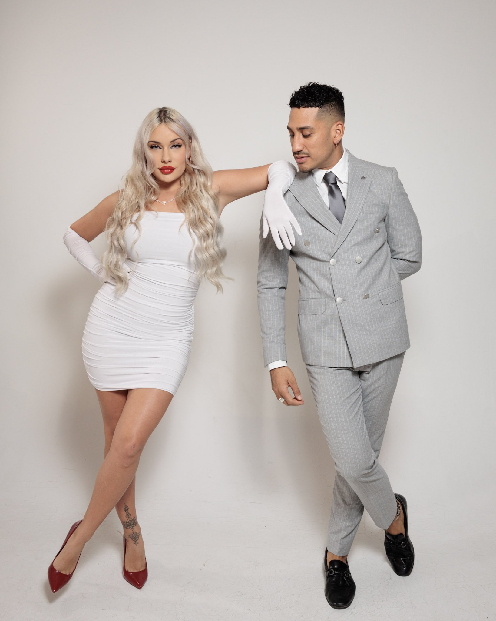 A woman in a tight white dress and red high heels leaning on a man in a gray suit with a white shirt and gray tie, both standing against a plain white wall.
