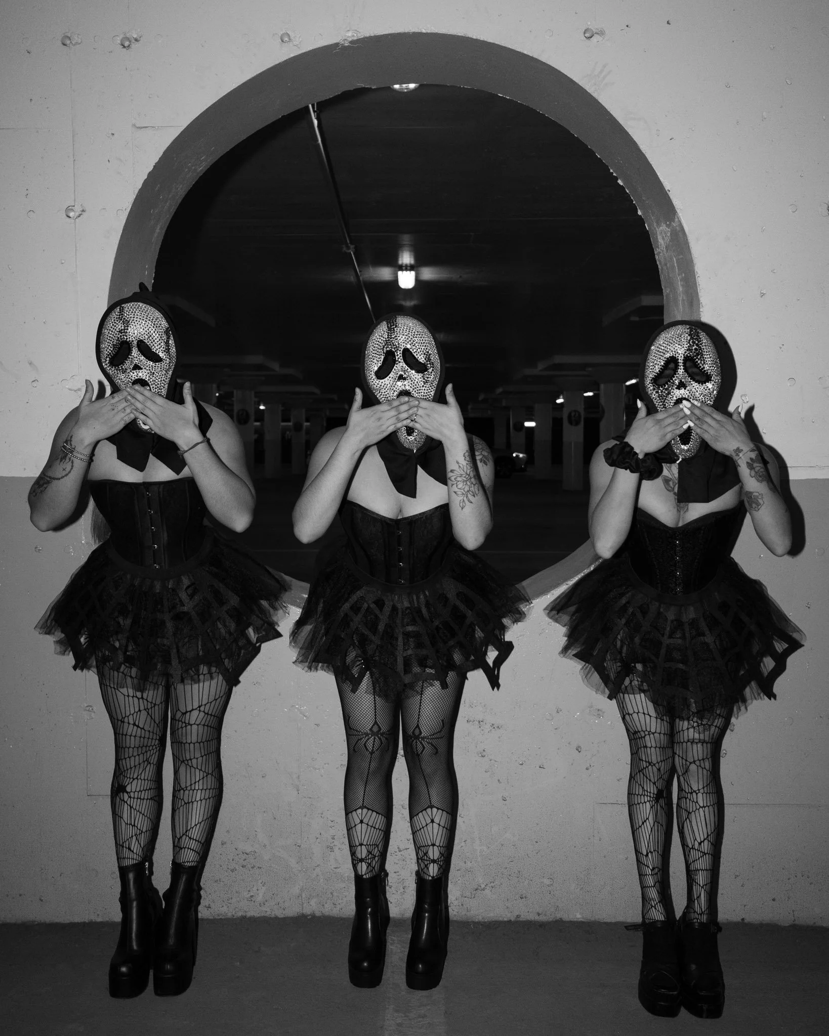 Three women wearing black corsets, black tutus, fishnet stockings, and platform boots, holding horror mask faces over their faces, standing in front of a mirror in a parking garage.