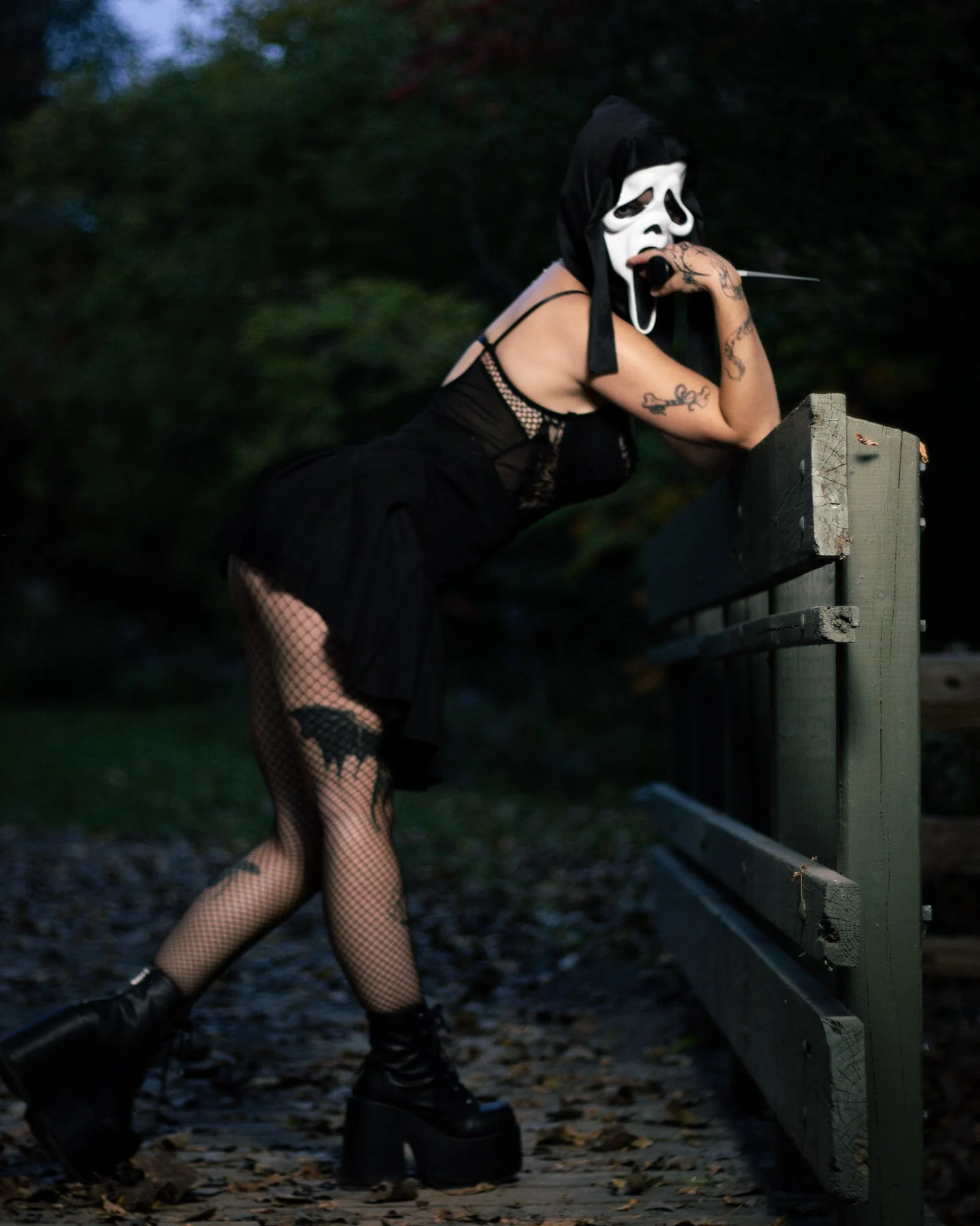 Person wearing a scream mask, black dress, fishnet stockings, and platform boots leaning on a wooden fence outdoors at dusk.