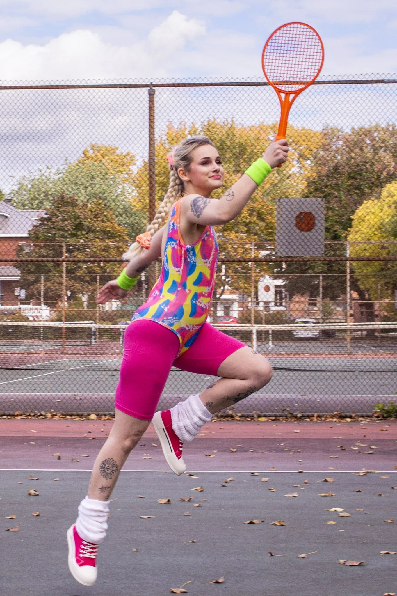 A young woman playing tennis on an outdoor court, with autumn trees and houses in the background.
