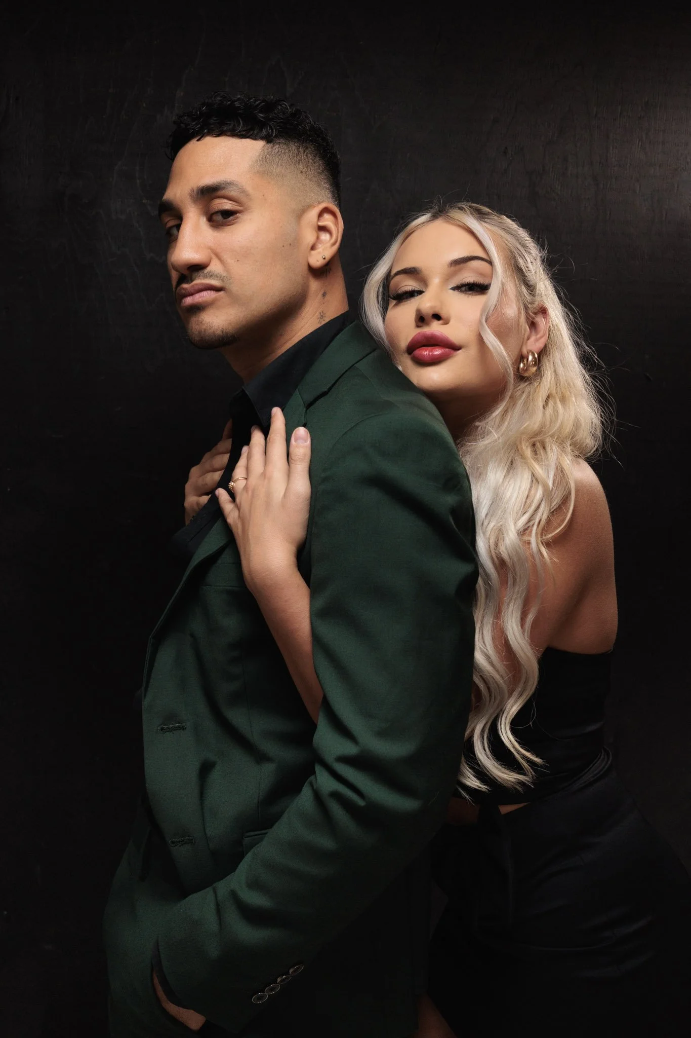 A man and woman posing against a black background, with the woman standing slightly behind and holding onto the man's shoulder.