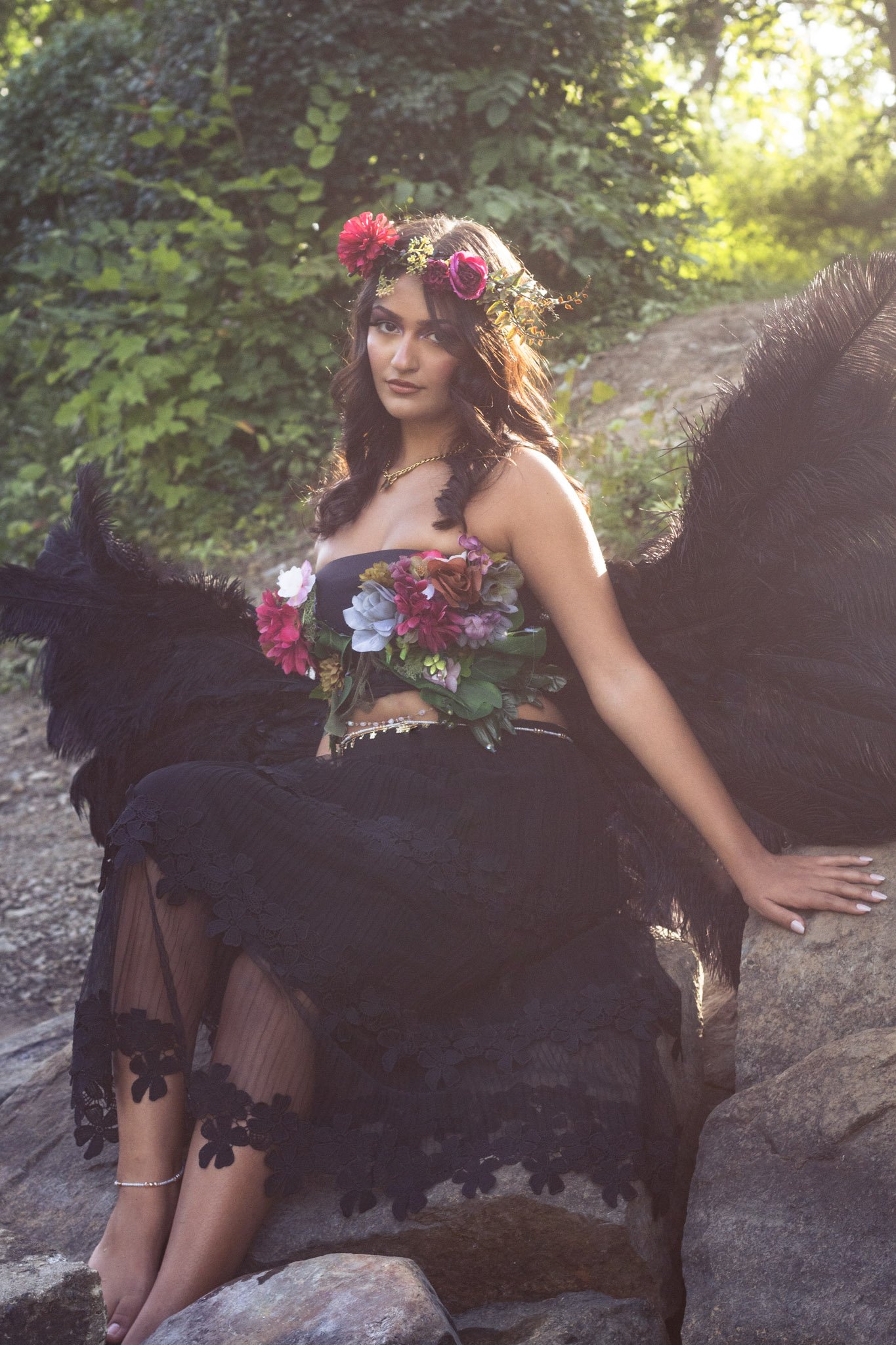 A woman sitting on rocks outdoors during sunset, dressed in a black lace skirt with floral appliqués, wearing a floral top and a flower crown, with large black feathered wings behind her.