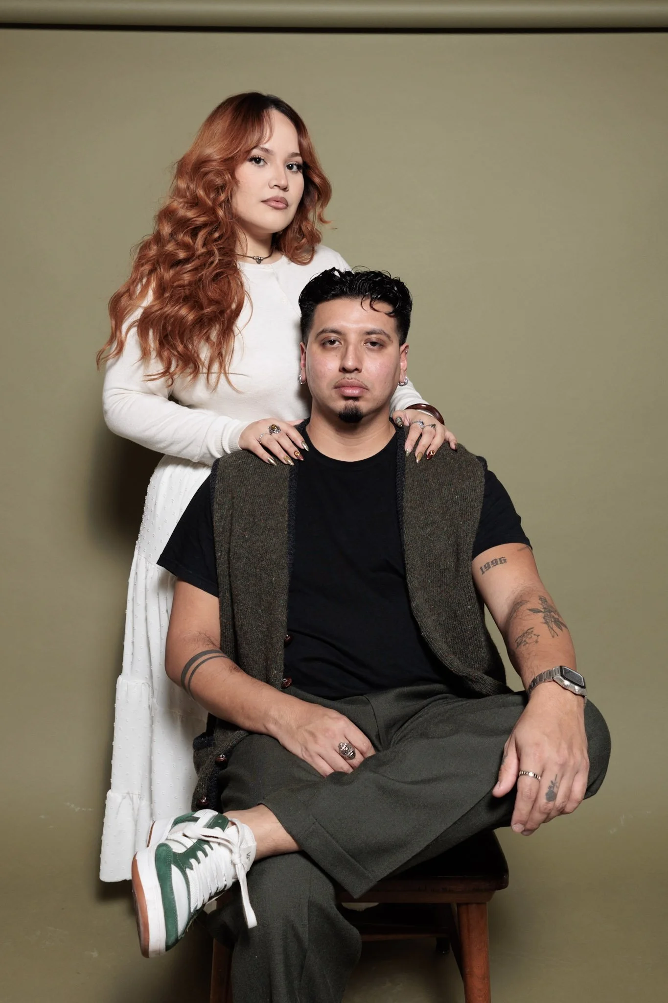 A woman with long, wavy red hair and a man with short black hair and tattoos, posing against a plain muted green background.