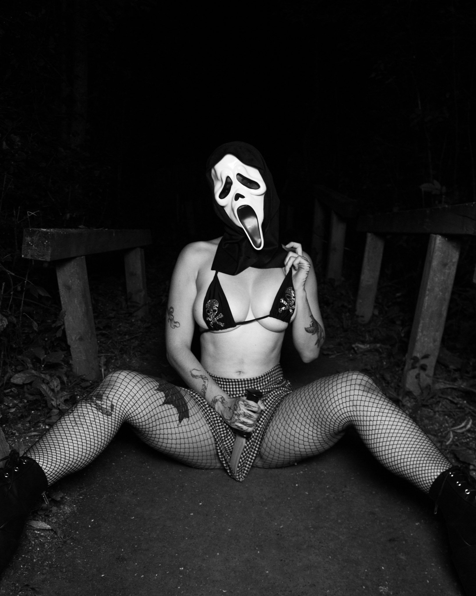 A person sitting on the ground wearing fishnet stockings, shorts, and a bra, holding a large knife, with a ghost mask and hood, in an outdoor wooded area at night.