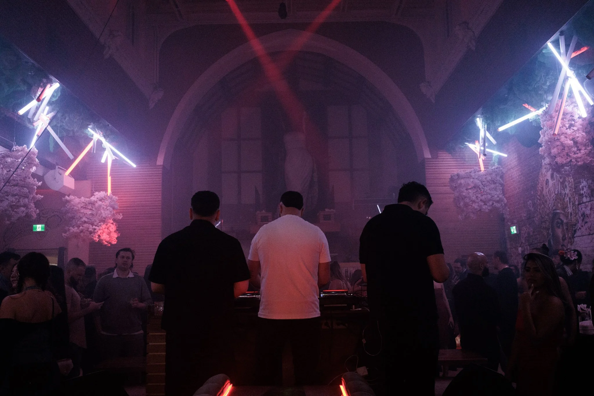 People dancing and socializing in a nightclub with pink and purple lighting, decorative trees, and a DJ in the center.