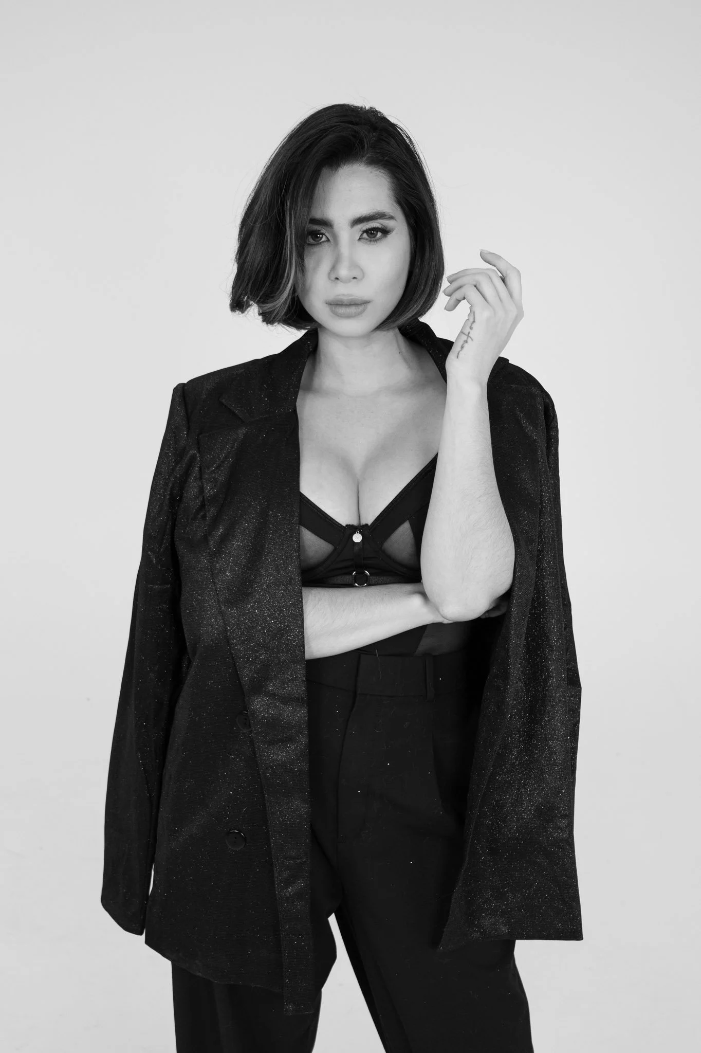 Black and white photo of a woman with short dark hair wearing a blazer, a revealing top, and high-waisted pants, standing with one hand near her face and the other crossed over her body.