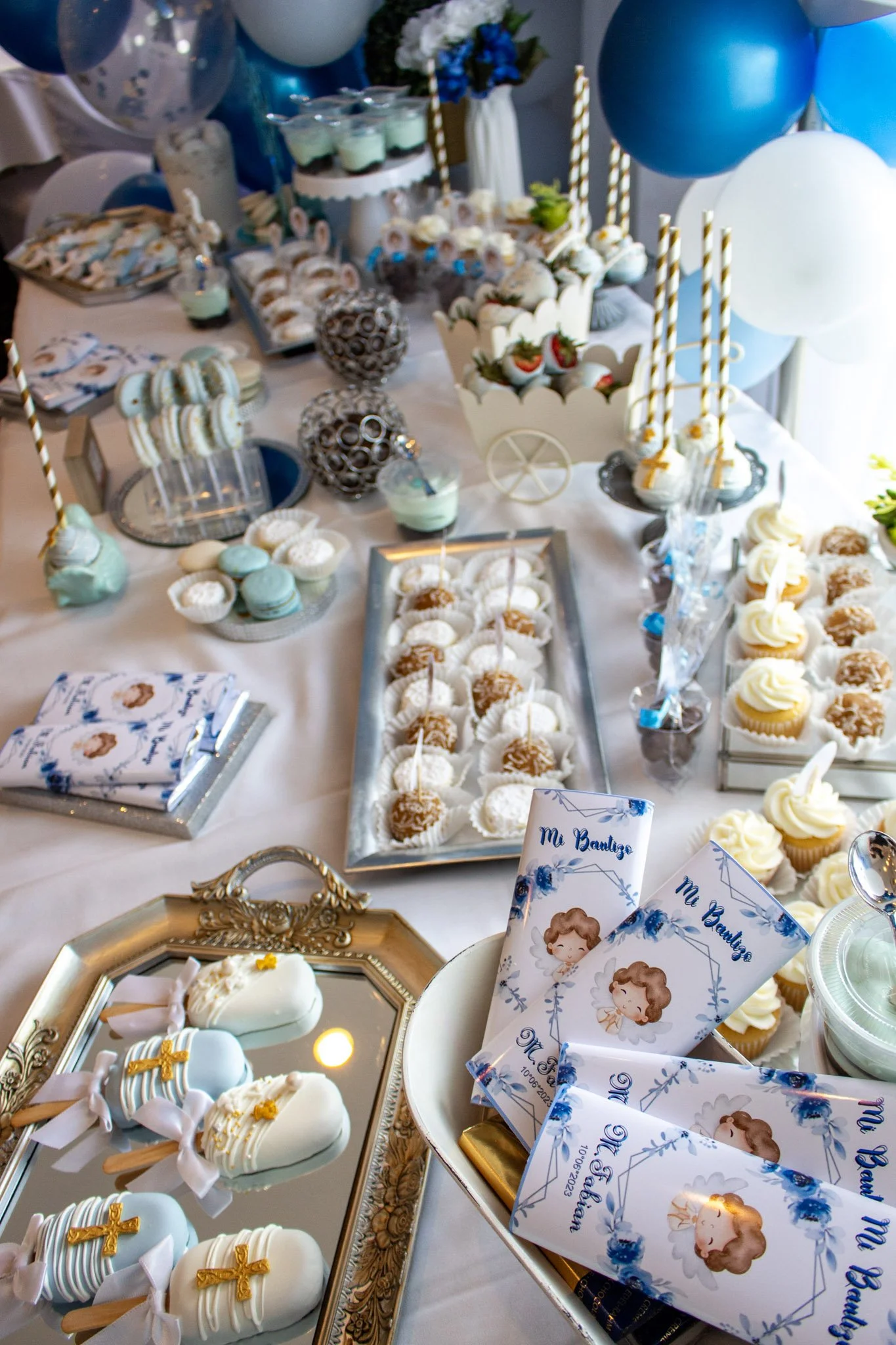A decorated table with blue and white themed treats and decorations for a celebration, including cupcakes, macarons, cake pops, and party favors, with blue, white, and silver balloons in the background.