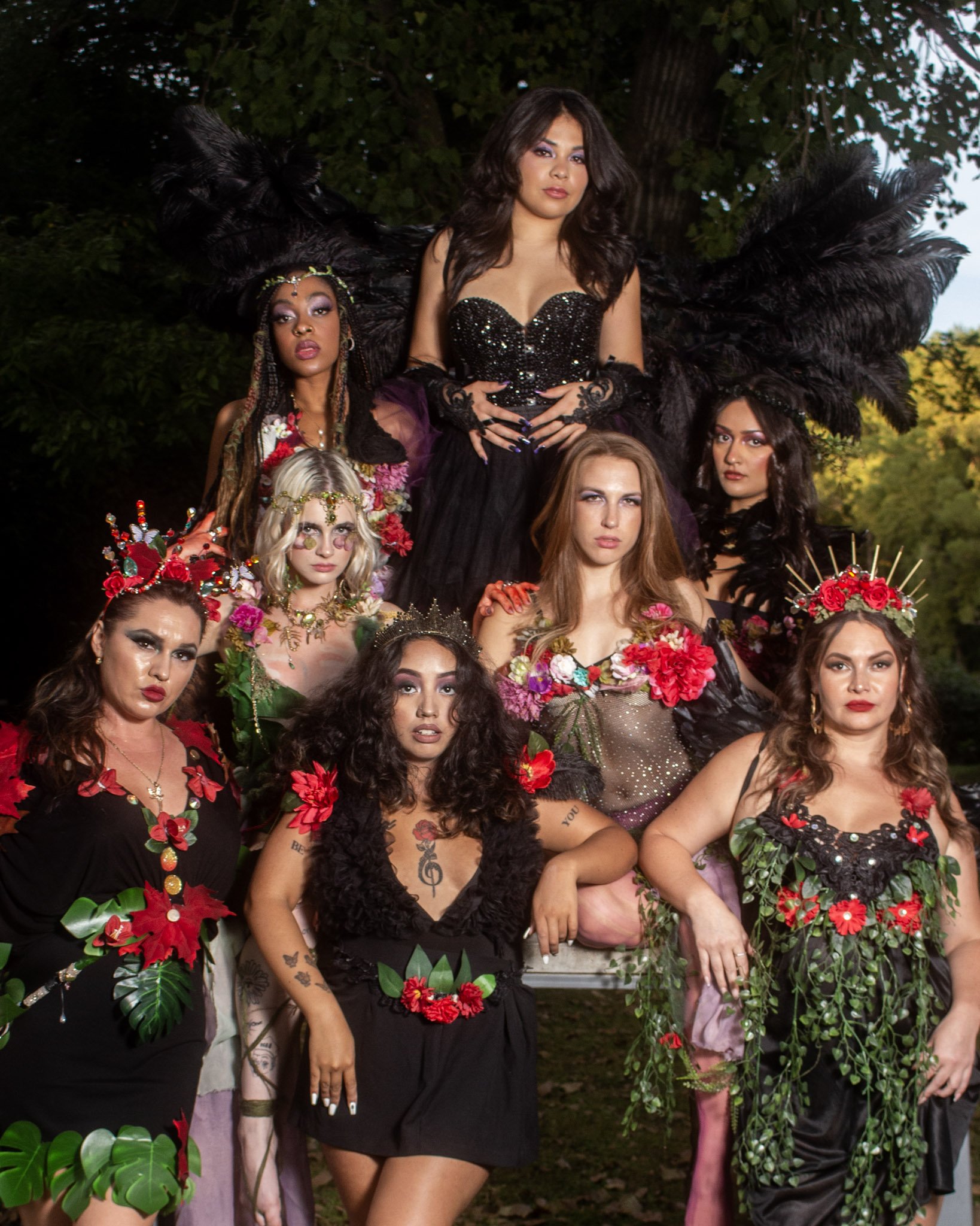 A group of diverse women dressed in elaborate, floral and black costumes with accessories, posing outdoors in front of trees.