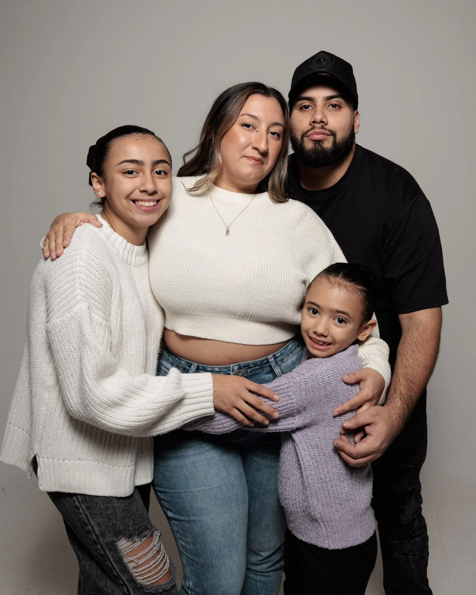 Family portrait of four people, two young girls, a woman and a man, standing together against a plain background, smiling and embracing each other.