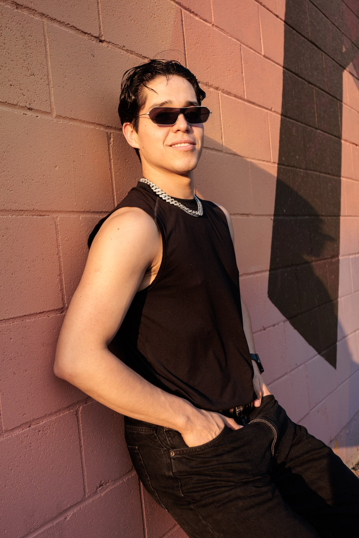 A person leaning against a pink brick wall during sunset, wearing sunglasses, a sleeveless black shirt, a silver chain necklace, and black pants.
