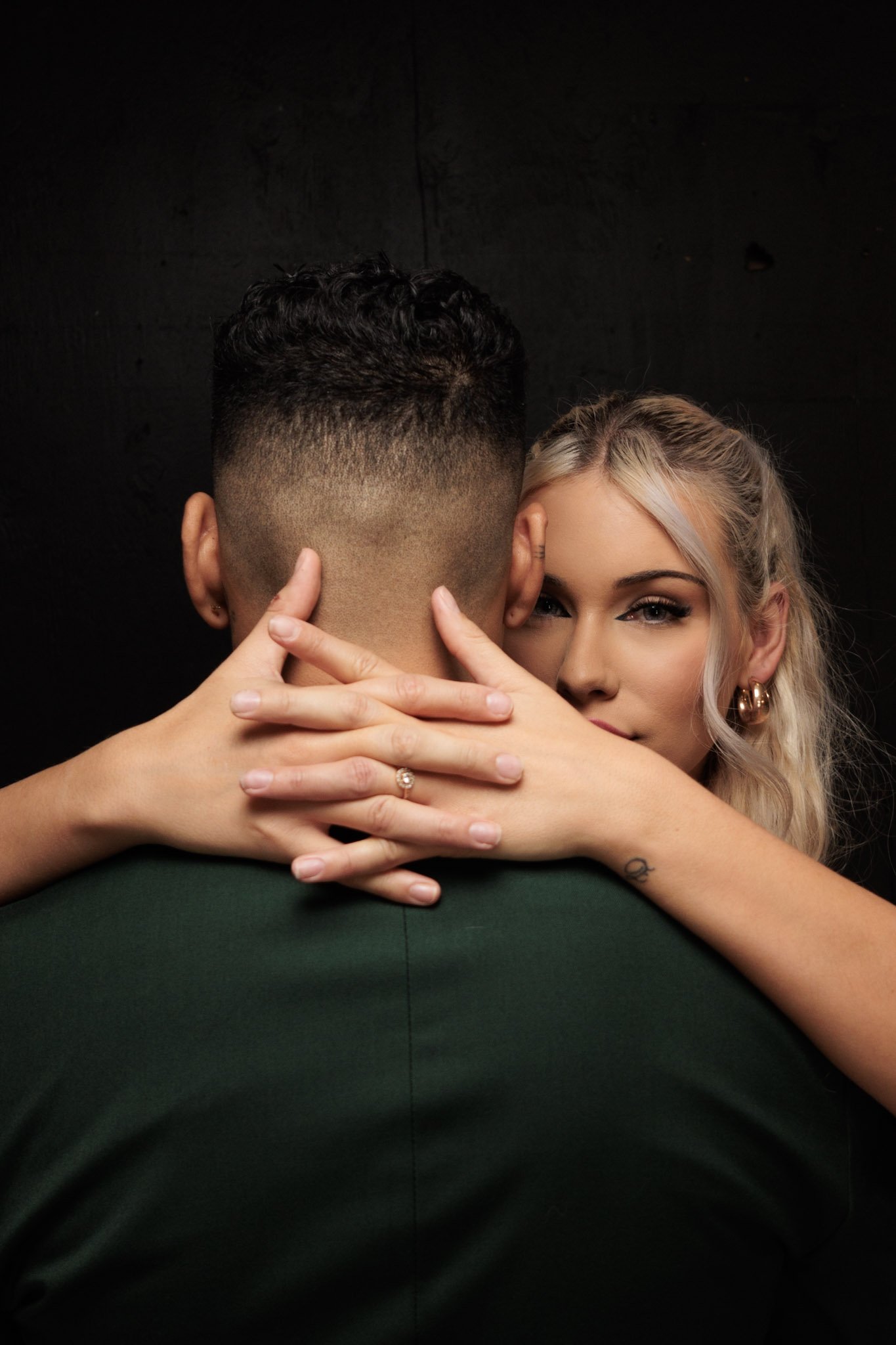 A woman with blonde hair hugging a man from behind, her face partially visible, against a dark background.