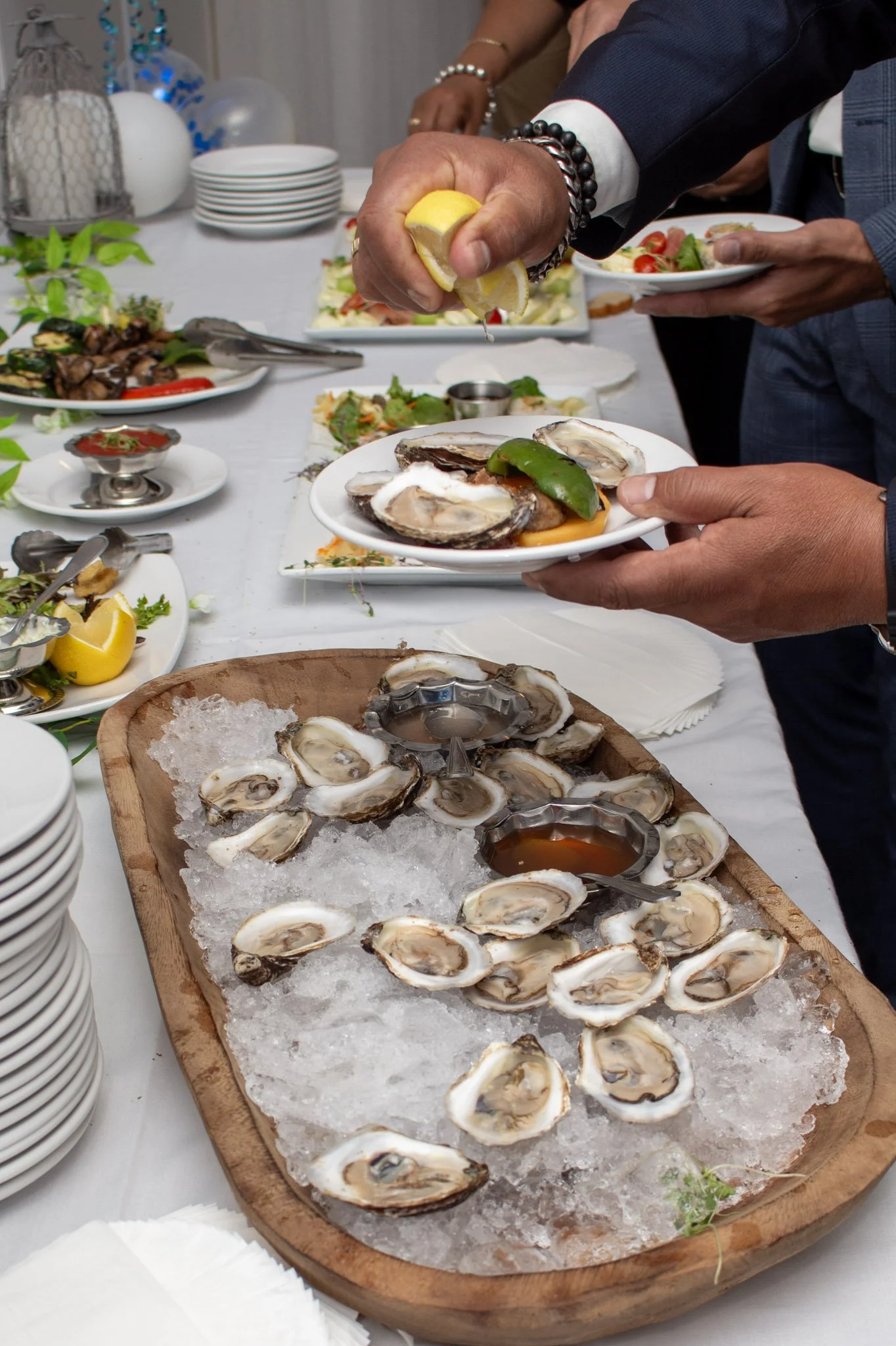 A seafood buffet with oysters on ice, someone squeezing lemon over oysters, and various plates of food on a white tablecloth at a social gathering.