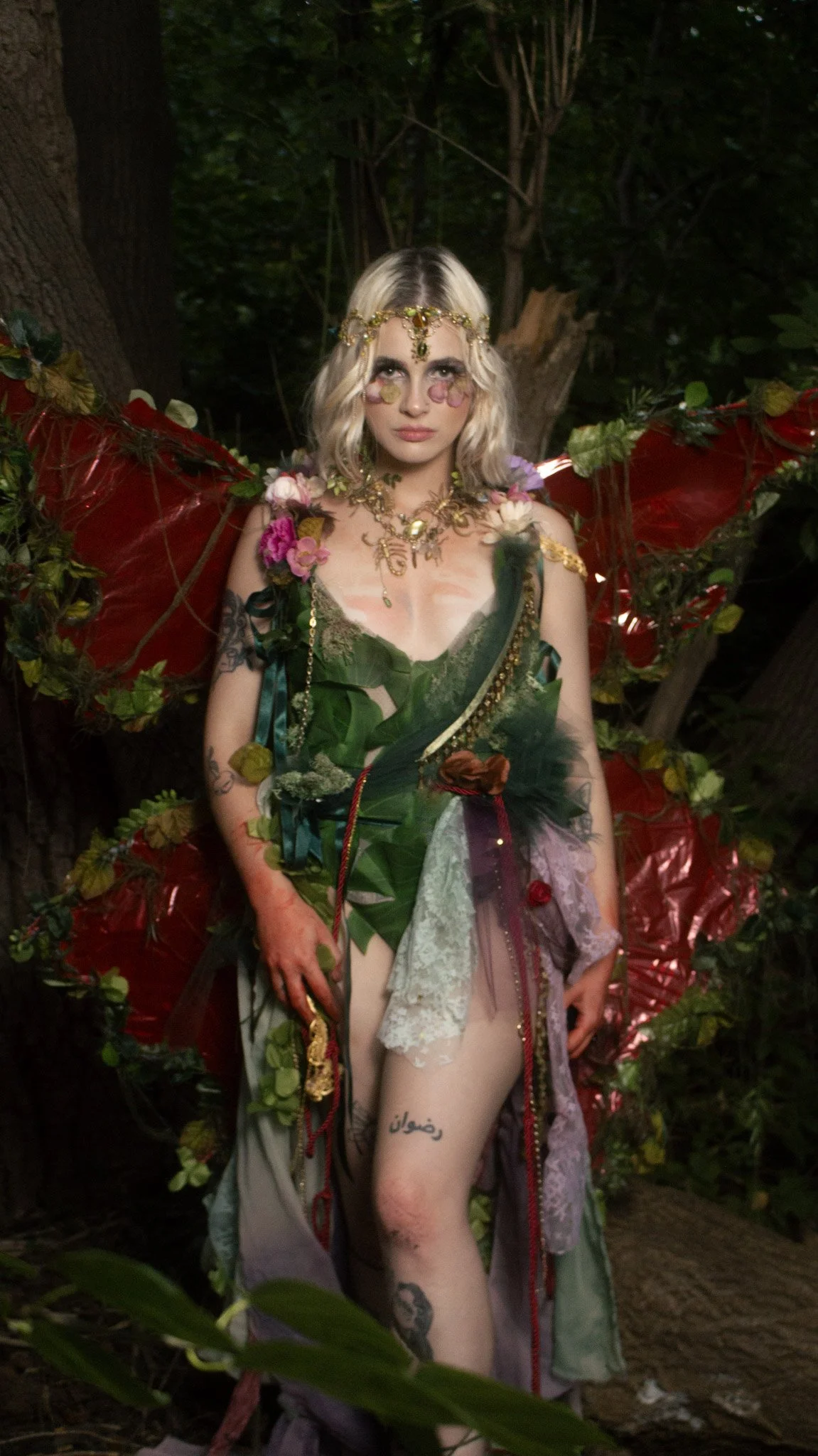 A woman dressed in a fantasy or fairy-inspired costume with floral and leaf details, wearing elaborate jewelry and a headpiece, standing outdoors among trees.