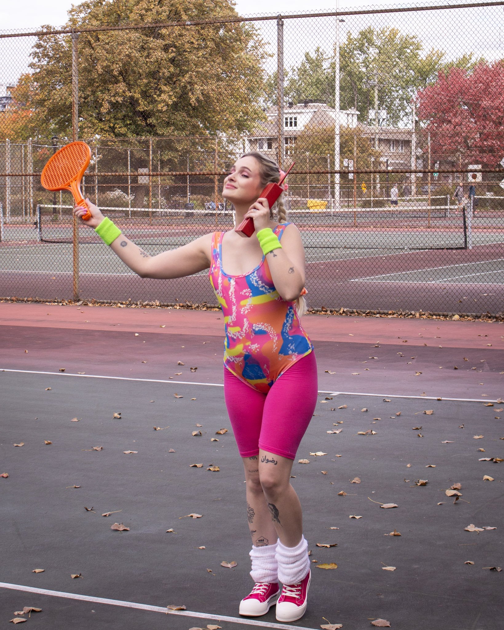 A young woman in colorful athletic wear holding a tennis racket and a red retro phone on a tennis court with fall trees in the background.