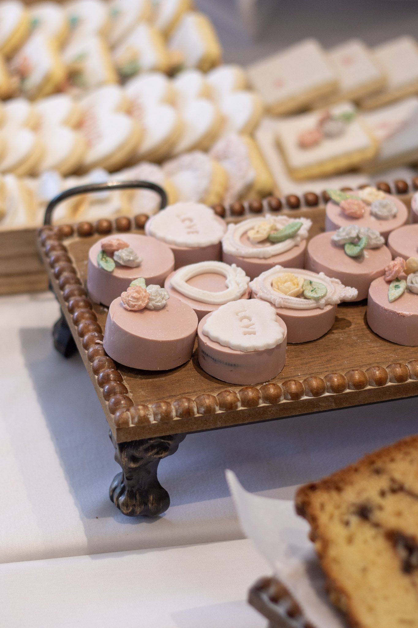 Decorative pink, white, and pastel-colored cookies with floral and heart designs on a wooden tray at a dessert display.