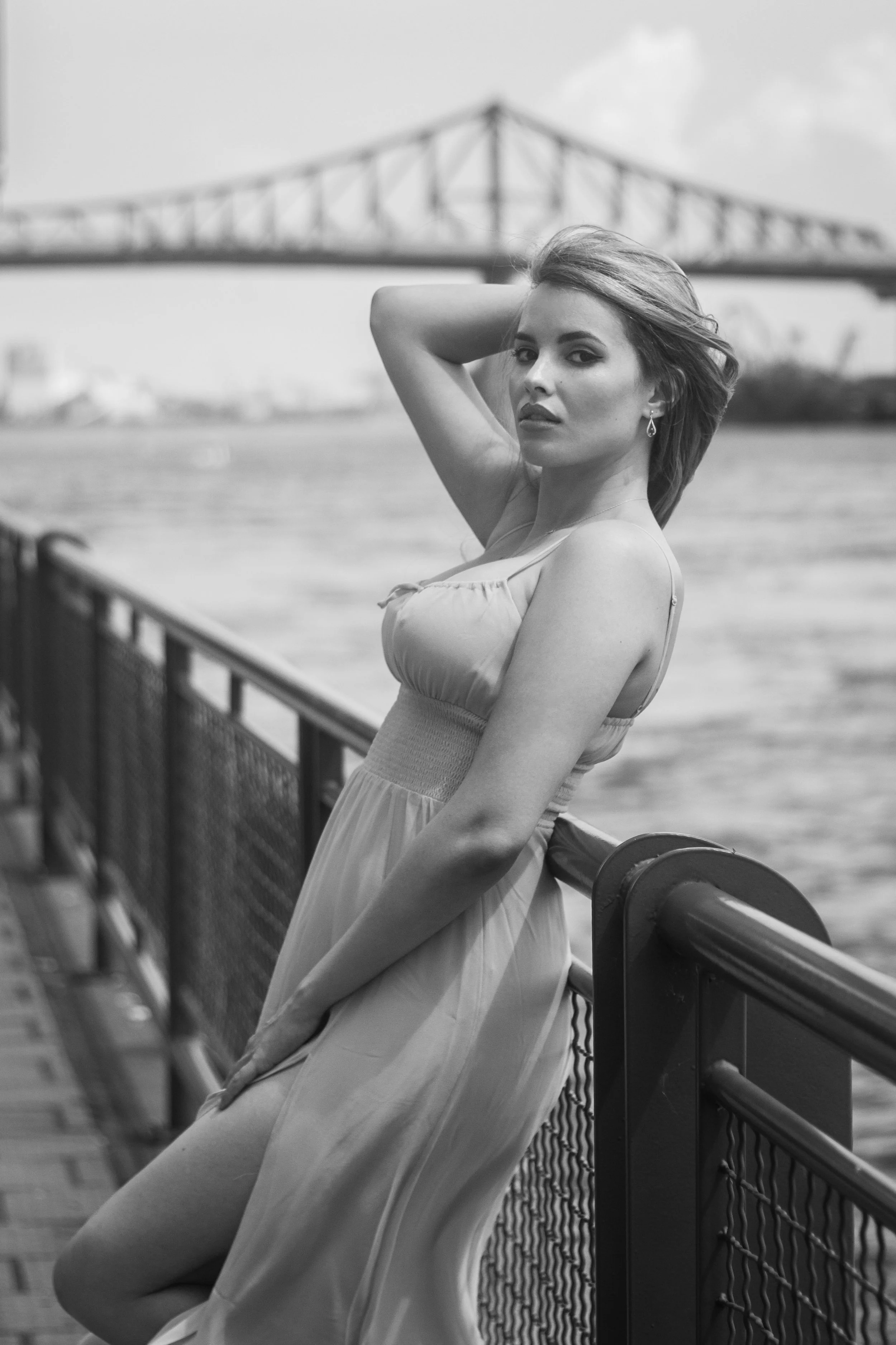 A woman in a dress leaning on a railing by the water with a bridge in the background. Outdoor photoshoot at le quaie de l'horloge in Montréal, Québec in Canada