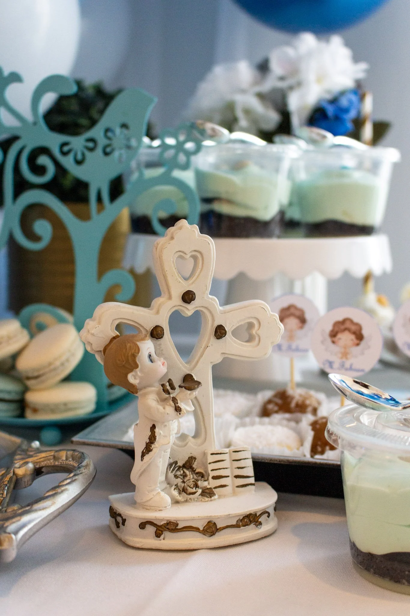 A decorative figurine of a boy praying next to a cross, part of a dessert table with macarons, cake, and balloons at a celebration.