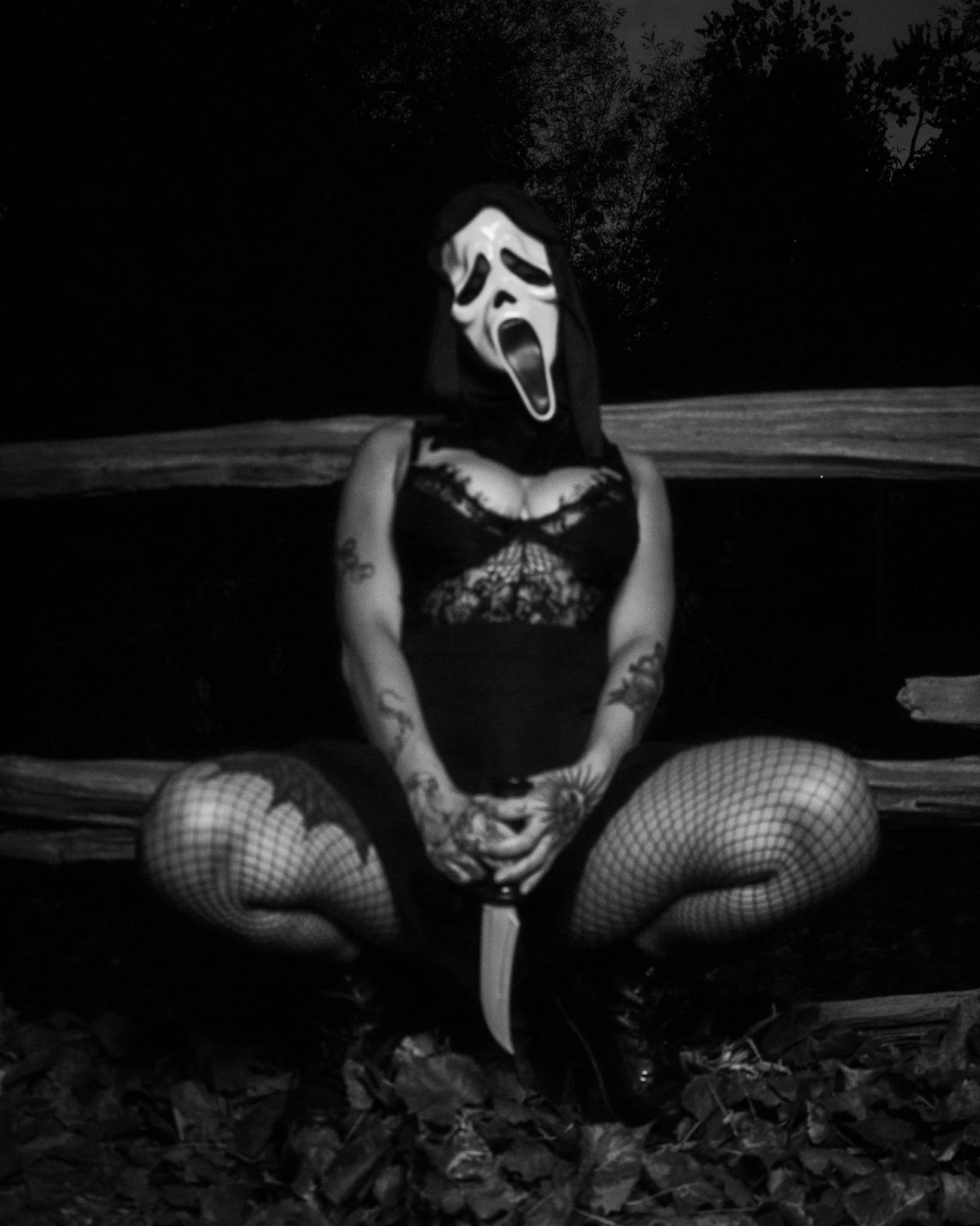 A person with tattoos on arms and legs wearing fishnet stockings, sitting cross-legged on the ground with a large knife in hands, wearing a mask resembling the Ghostface character from horror movies, outdoors at night.