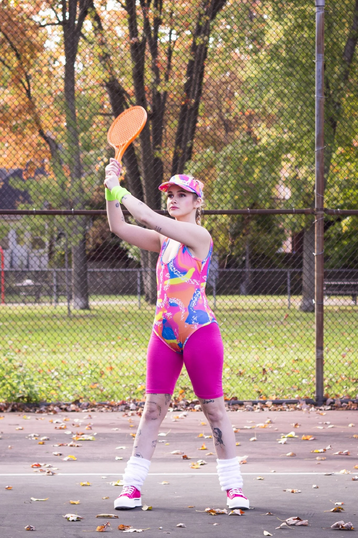 A young woman in colorful athletic wear preparing to serve a tennis ball on an outdoor court, with a chain-link fence and autumn trees in the background.