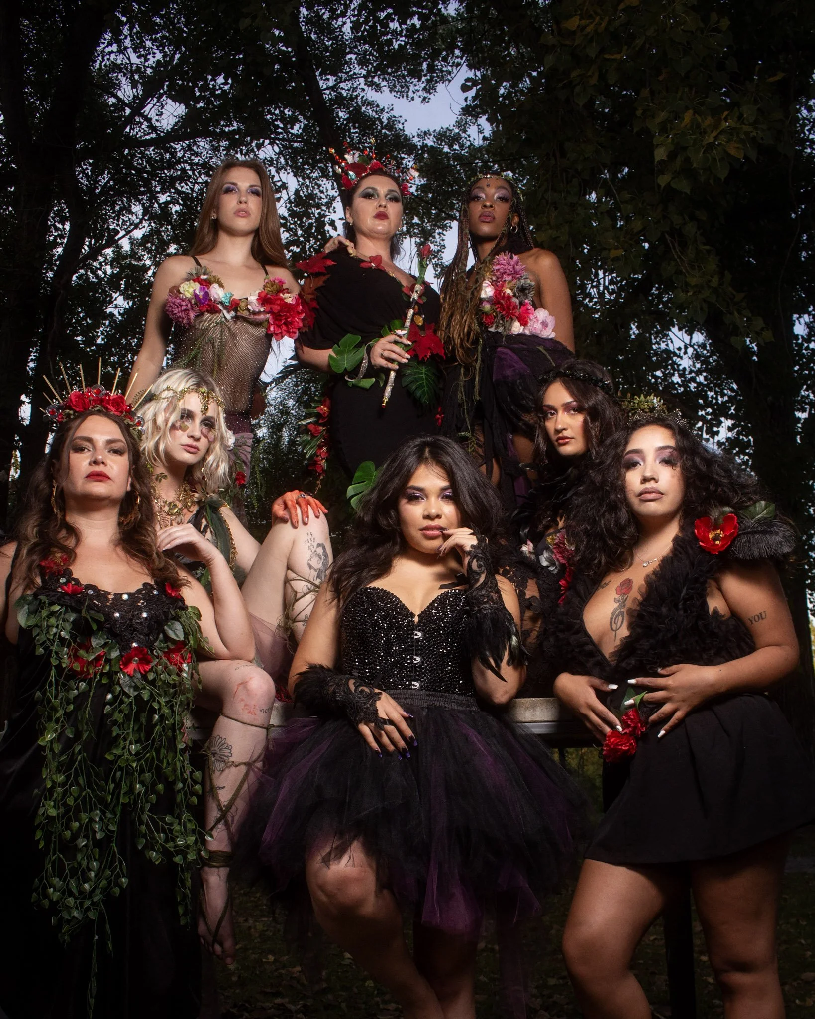 A diverse group of nine women dressed in elaborate, dark, floral-themed costumes posing outdoors against a backdrop of trees at dusk.