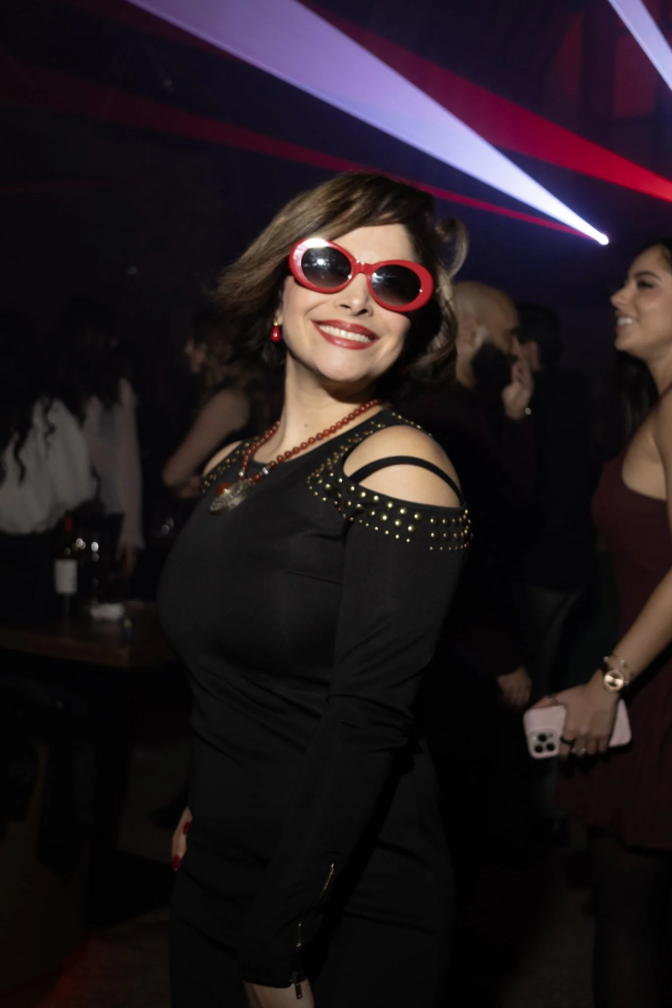 A woman with shoulder-length dark hair wearing large red sunglasses, a black dress with shoulder cut-outs and gold studs, red earrings, and a red necklace, smiling at a party with colorful lighting and other guests in the background.