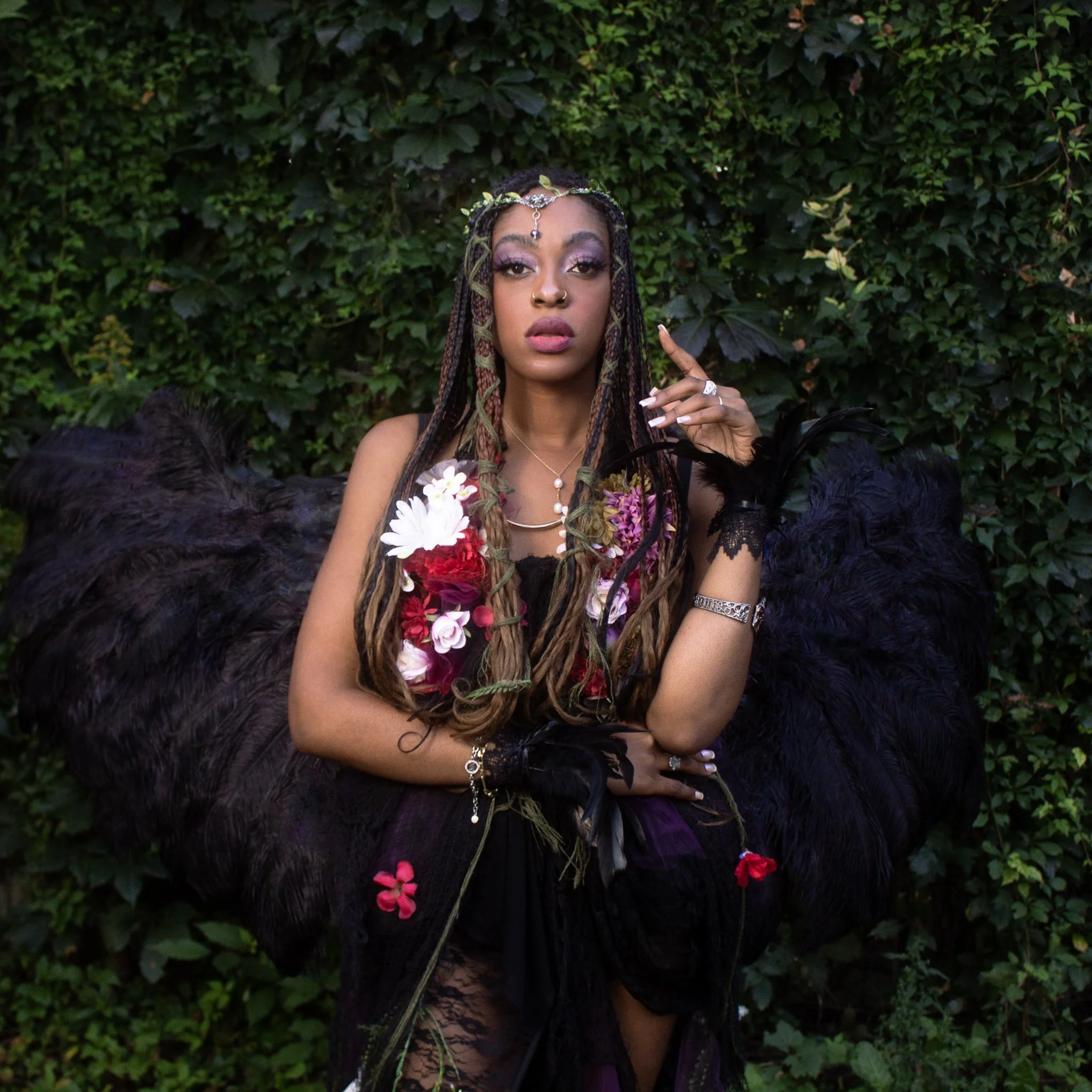 A woman dressed in a fantasy-inspired costume with black feathered wings, floral adornments on her dress, and long braided hair stands against a lush, green leafy background. She has jewelry, makeup, and poses with one finger raised.